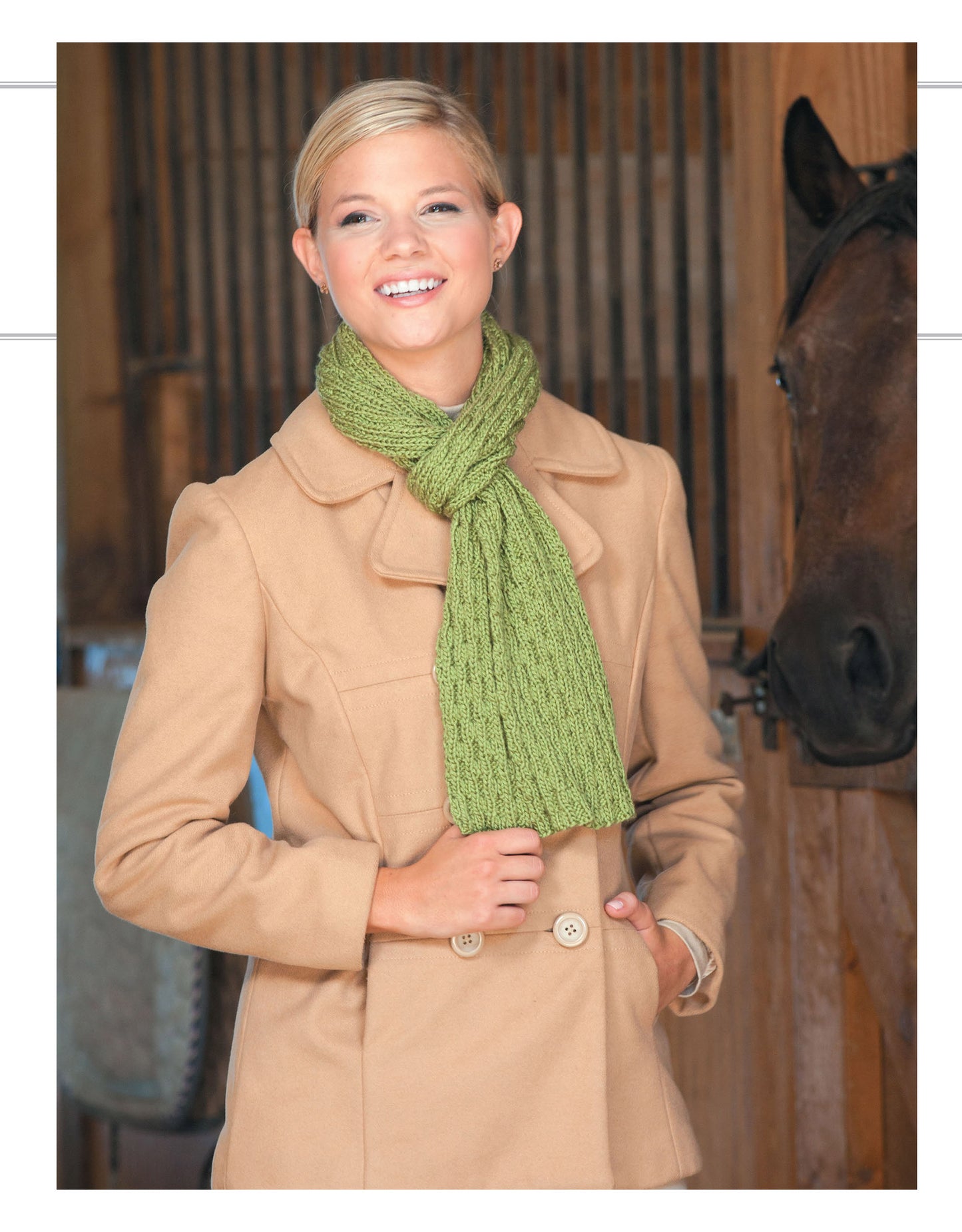 A smiling woman wears a green rib-knit scarf from Cozy Scarves Knitting by Leisure Arts and a tan coat, standing indoors beside a brown horse with wooden stalls in the background.