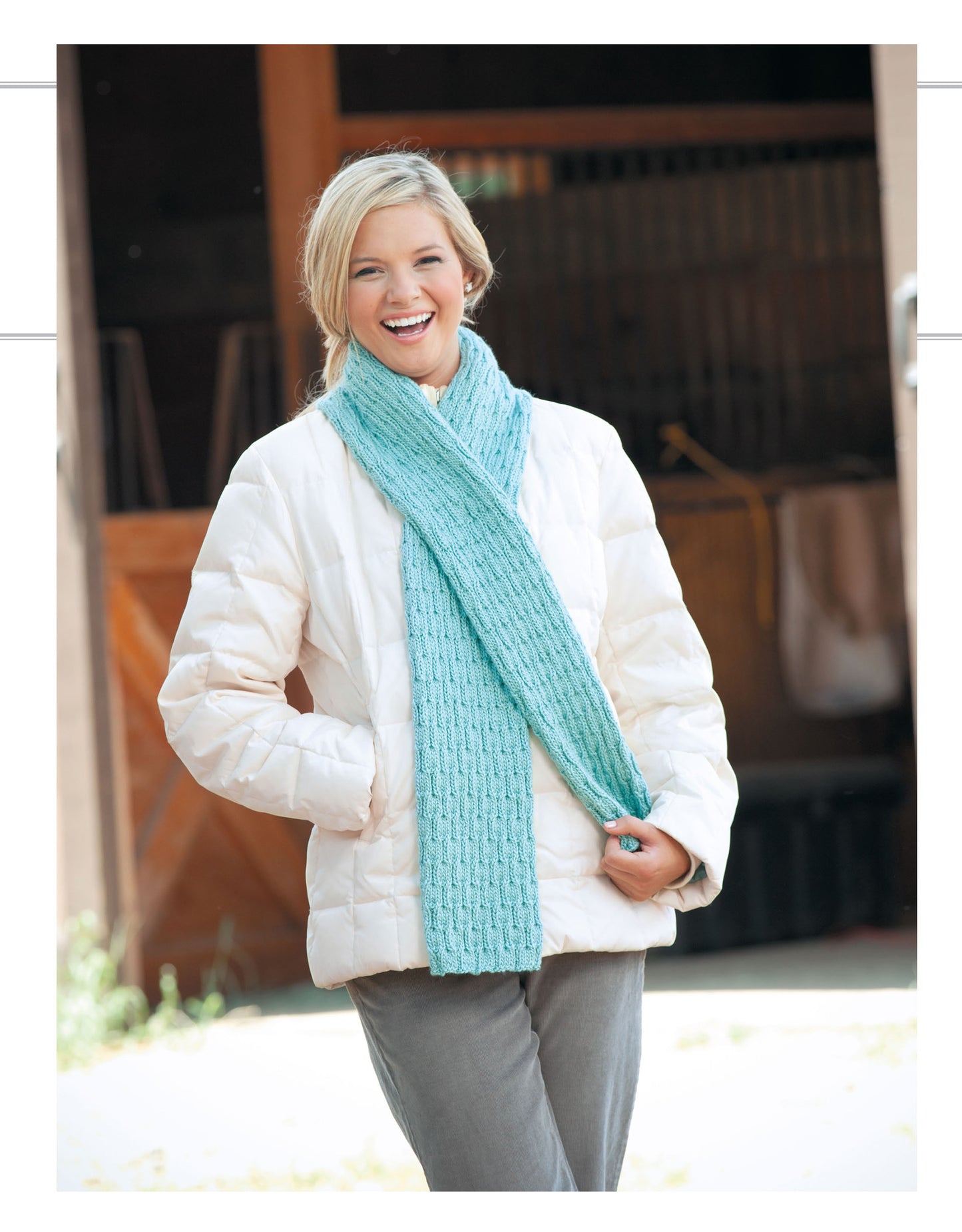 A smiling woman in a white puffer jacket and gray pants stands outdoors by a rustic wooden structure, wearing a light blue scarf made with Cozy Scarves Knitting patterns from Leisure Arts.