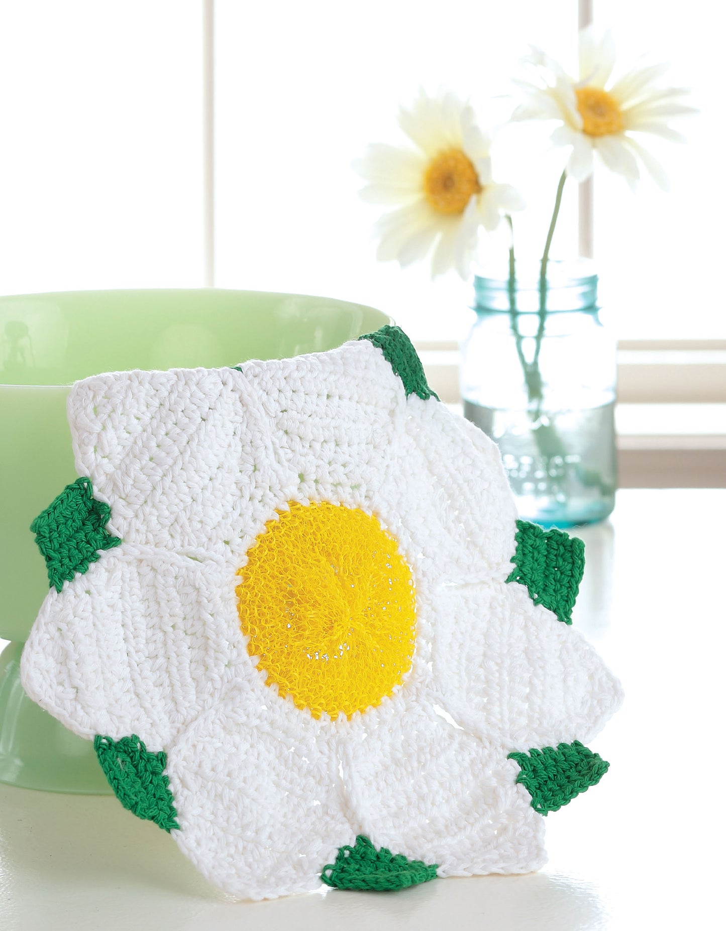 A vibrant crocheted dishcloth from Leisure Arts' "Quick & Colorful Dishcloths Designs to Brighten Any Kitchen" digital download is displayed near a pastel bowl, with real daisies in a blue mason jar set by a sunny window.