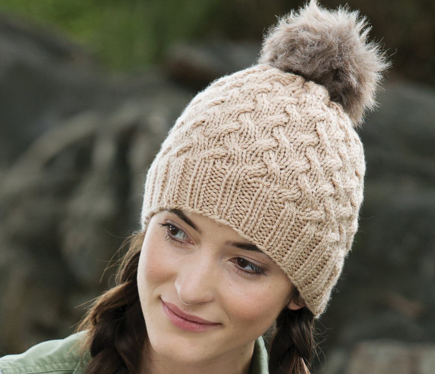 A young woman with brown hair in braids wears a beige knitted hat with a fluffy pom-pom, created from Leisure Arts' Casual Weekend Knits—perfect for beginners—while sitting outdoors among blurred rocks and greenery.