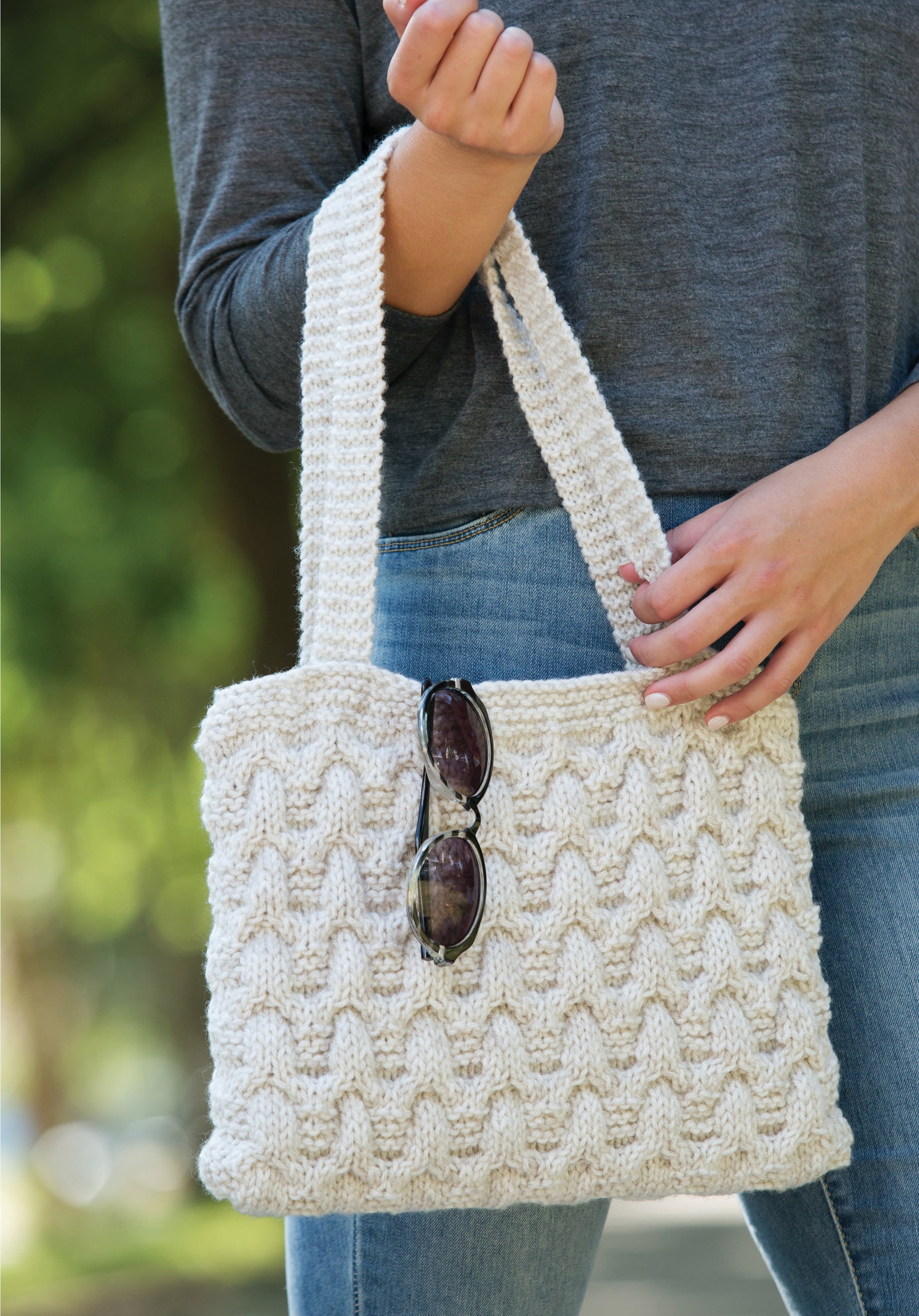A person in a gray shirt and blue jeans holds a textured, cream-colored knit handbag from Casual Weekend Knits by Leisure Arts—perfect for beginner or new wearable patterns. Sunglasses hang on the bag, with greenery in the outdoor background.