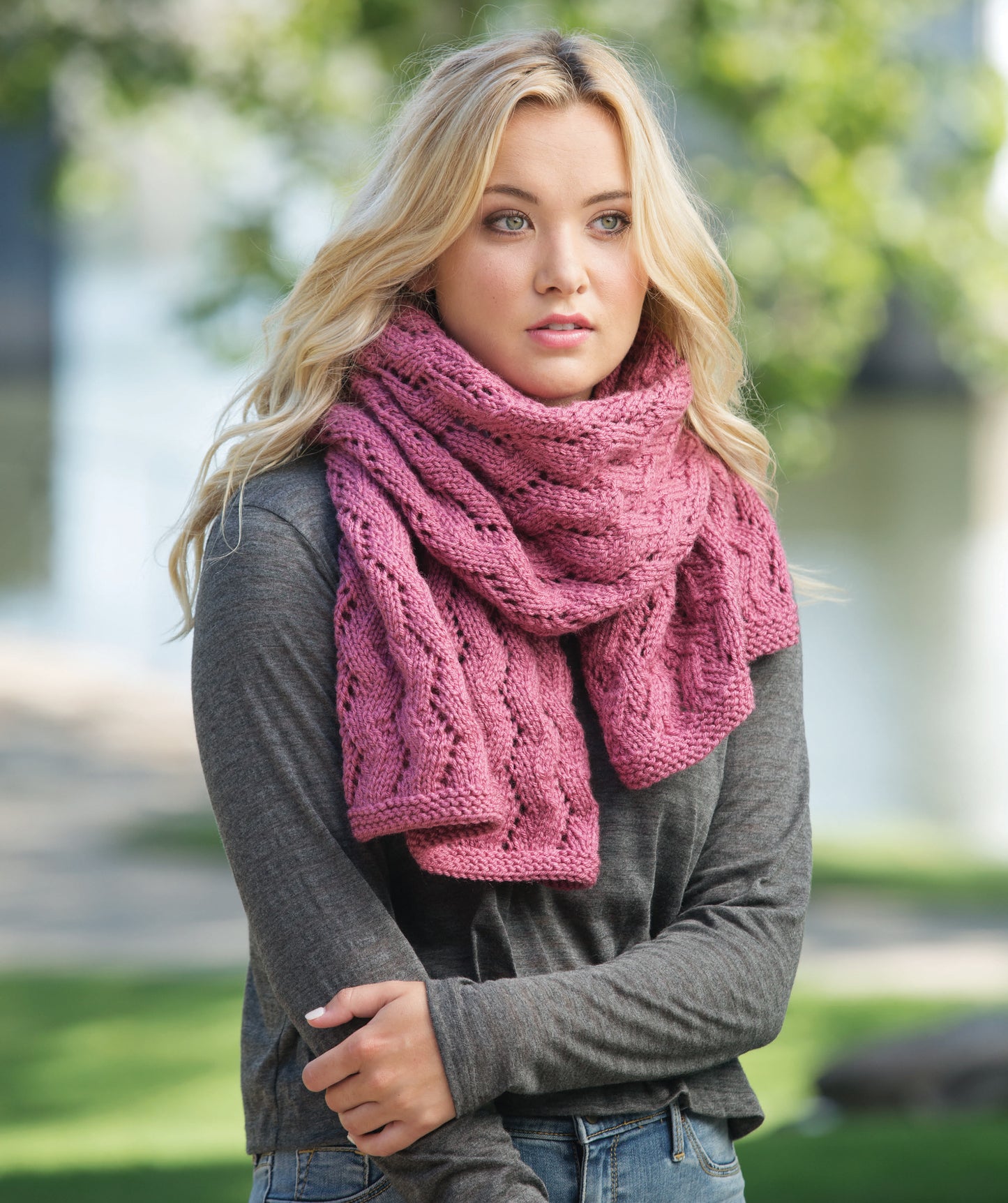 A woman with long blonde hair models a chunky pink scarf from "Casual Weekend Knits" by Leisure Arts, paired with a gray long-sleeve shirt, standing outdoors amid park greenery and a softly blurred background.