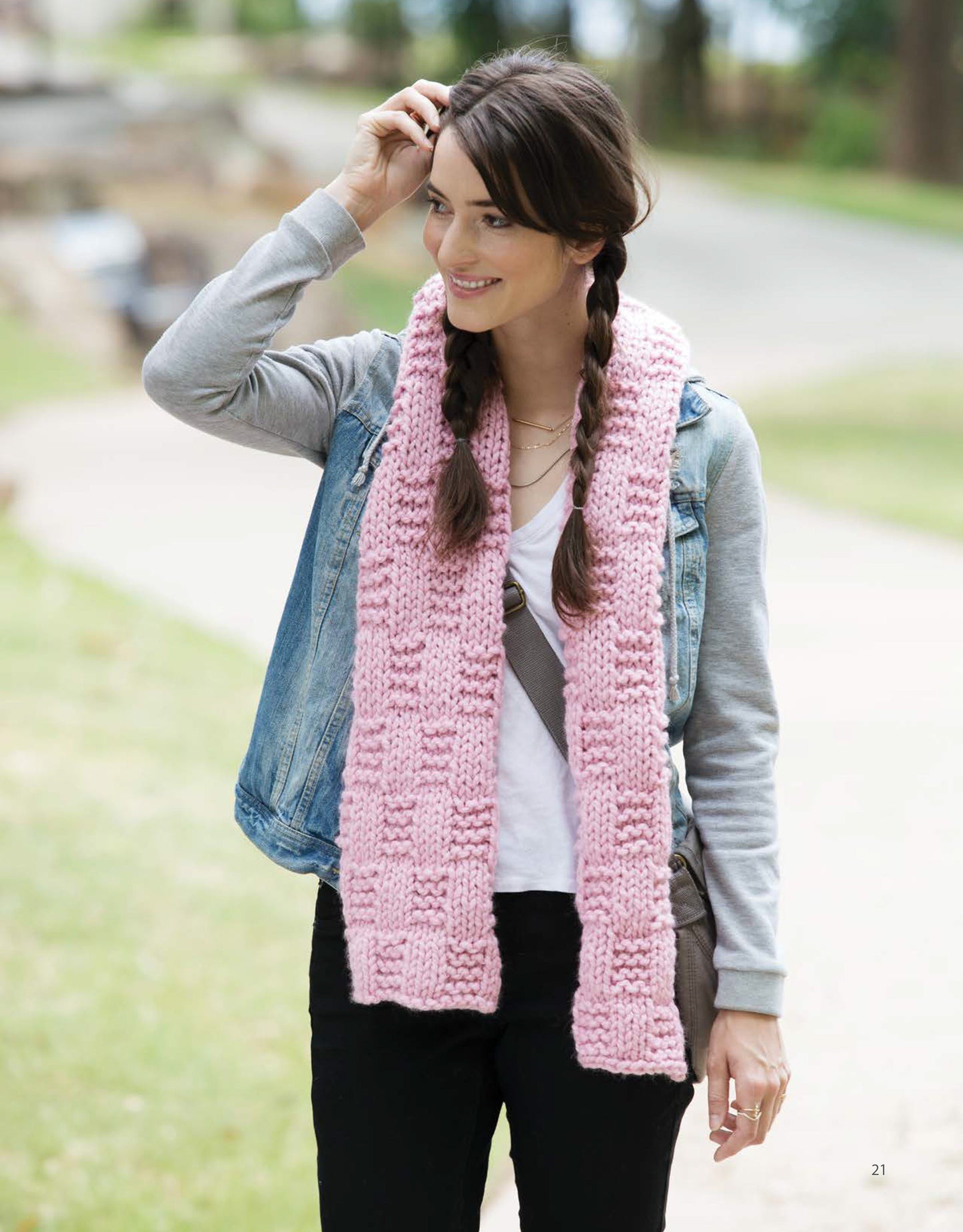 A woman with braided hair smiles while walking outdoors in a denim vest, gray hoodie, black pants, and a pink chunky knit scarf. Her cozy accessory was made using "Casual Weekend Knits" by Leisure Arts—a great pick for beginner knitting patterns.