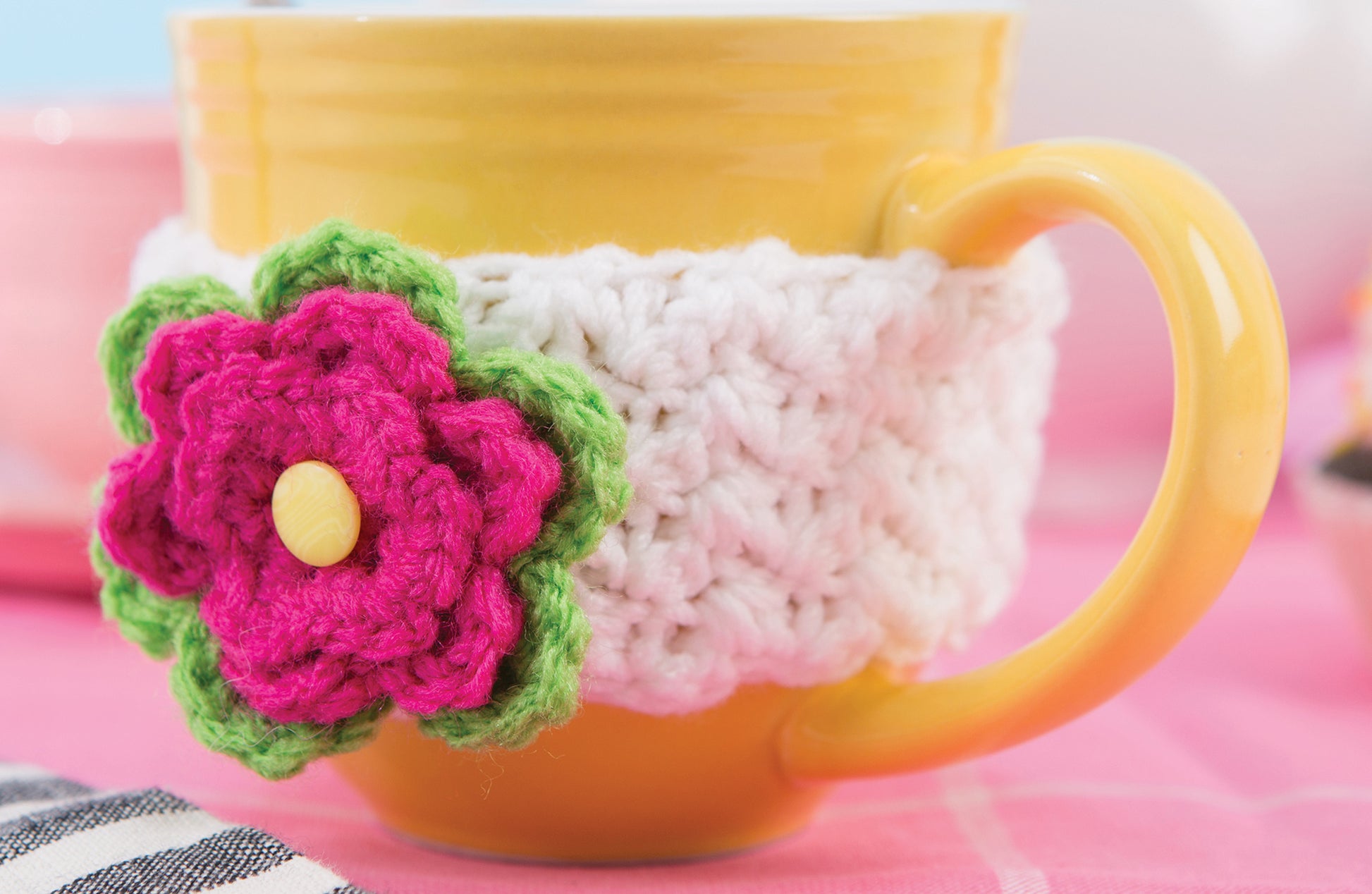 A yellow mug with a white crocheted cozy, decorated with a pink and green flower, rests on a pink checkered surface—an inspiring showcase for Leisure Arts’ “Happy Crochet—13 Fun Projects That Are Sure to Brighten Your Day.”.