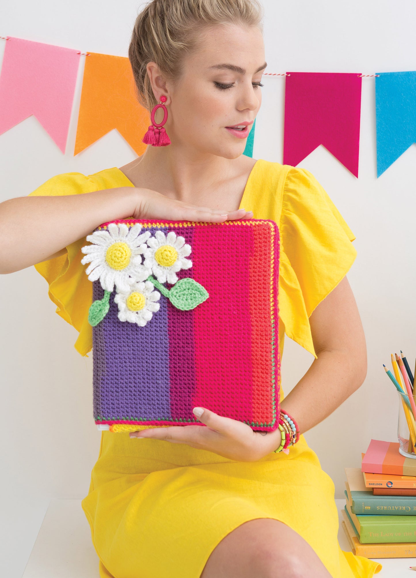 A woman in a yellow dress holds the "Happy Crochet—13 Fun Projects That Are Sure to Brighten Your Day" notebook by Leisure Arts, featuring a crocheted white daisy cover. She wears pink earrings and sits before bright pennant banners.
