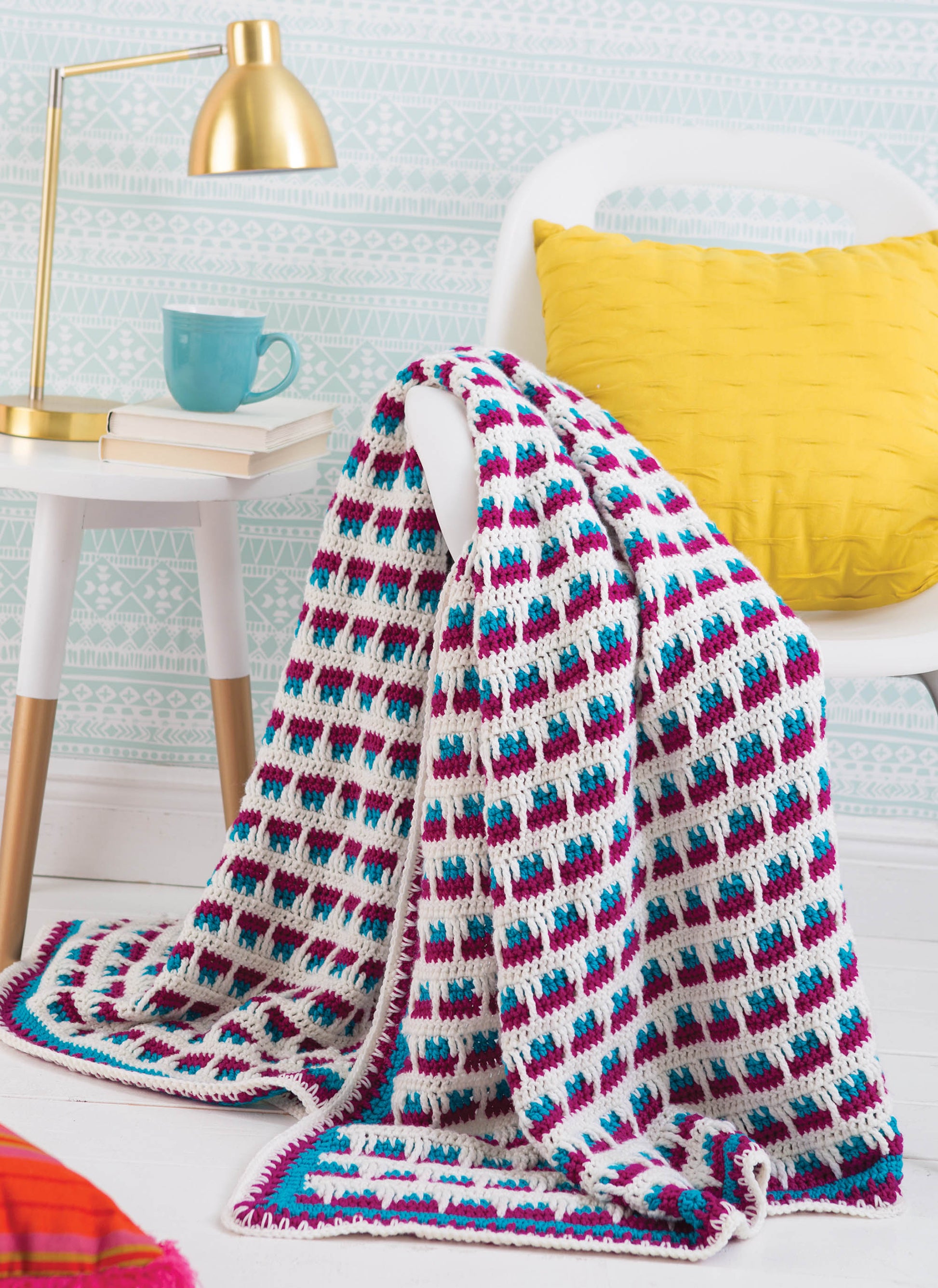Leisure Arts' "Happy Crochet—13 Fun Projects That Are Sure to Brighten Your Day" features a vibrant geometric blanket in white, purple, pink, and blue, perfect as an easy project to add color and cheer to any room.