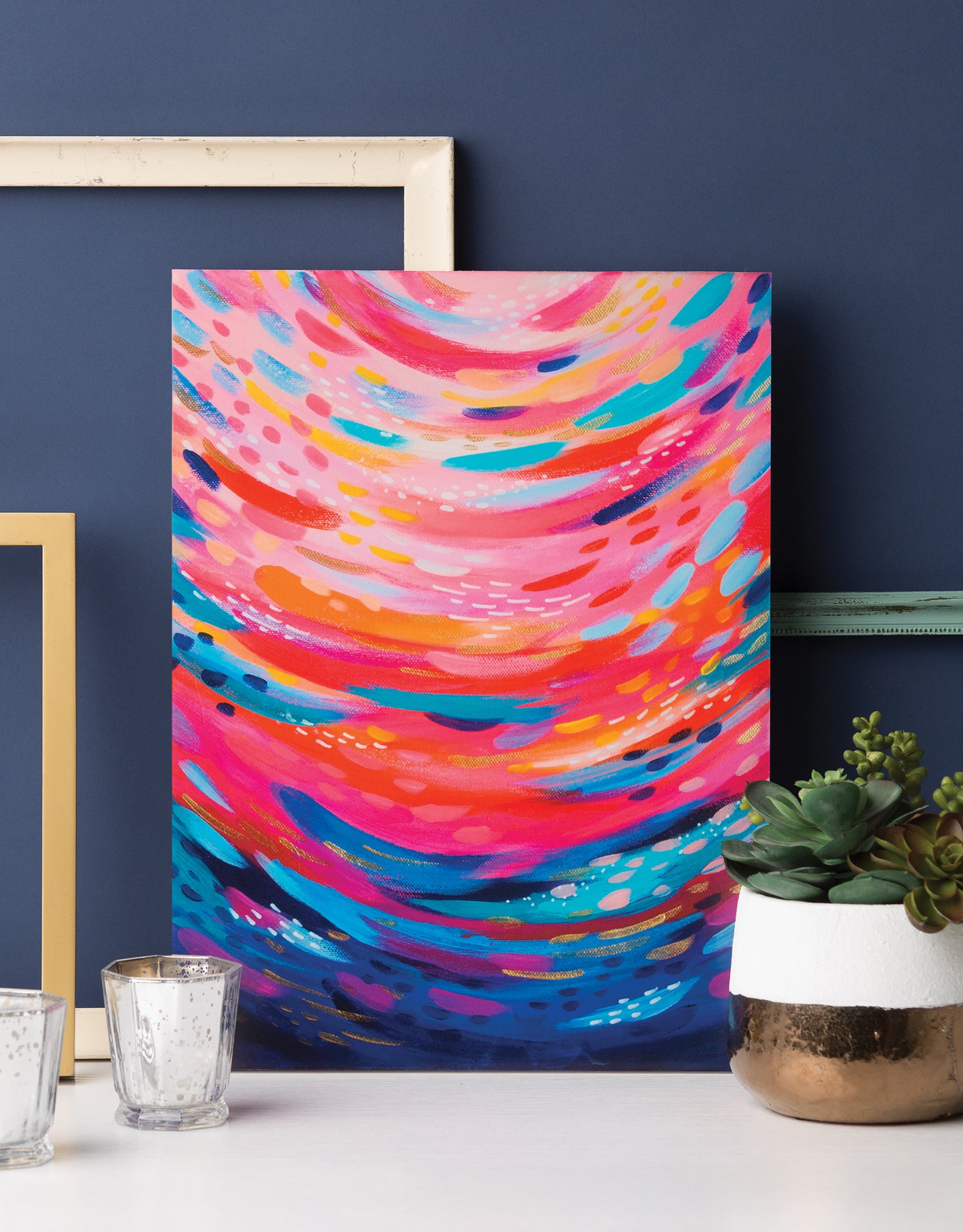 The Leisure Arts "Happy Abstracts—Fearless Painting for True Beginners" canvas art, featuring swirling pink, blue, orange, and white patterns, is showcased on a dark blue wall beside empty frames and a small potted succulent.