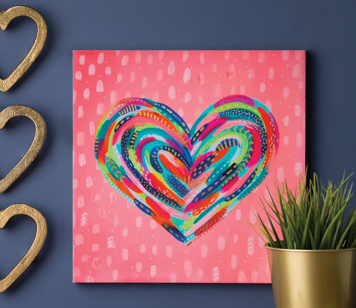 “Happy Abstracts—Fearless Painting for True Beginners” by Leisure Arts is displayed on a blue wall, featuring colorful heart artwork beside gold heart decorations and a green plant in a gold pot.
