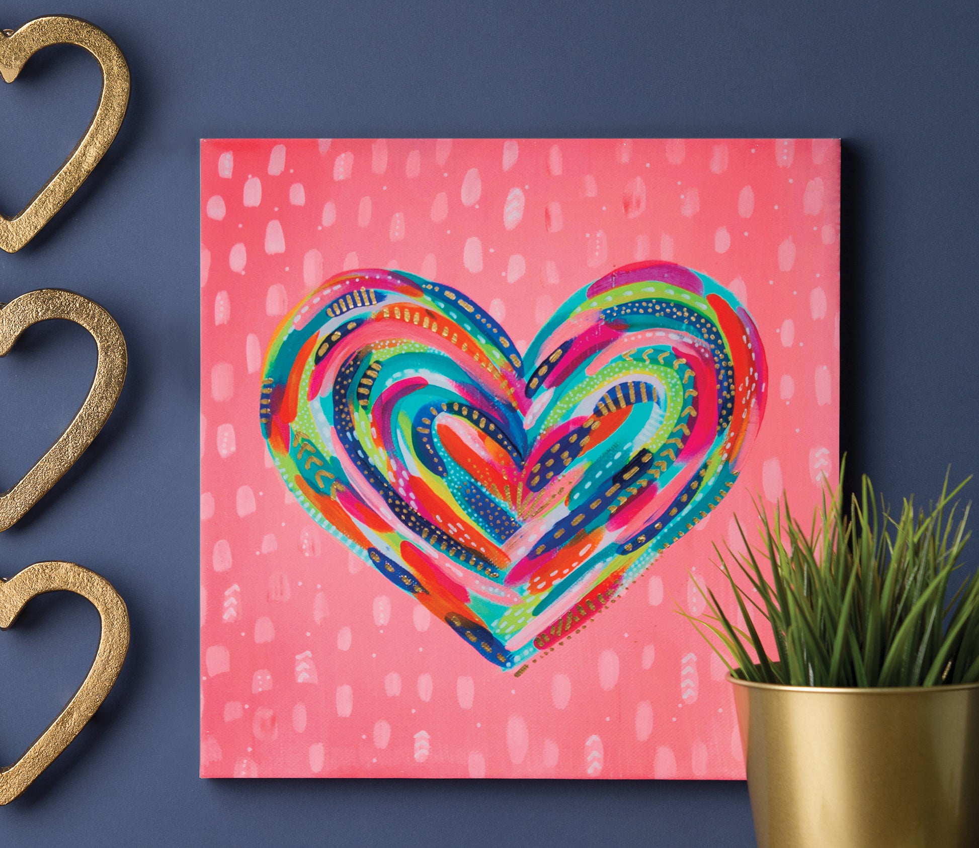 “Happy Abstracts—Fearless Painting for True Beginners” by Leisure Arts is displayed on a blue wall, featuring colorful heart artwork beside gold heart decorations and a green plant in a gold pot.