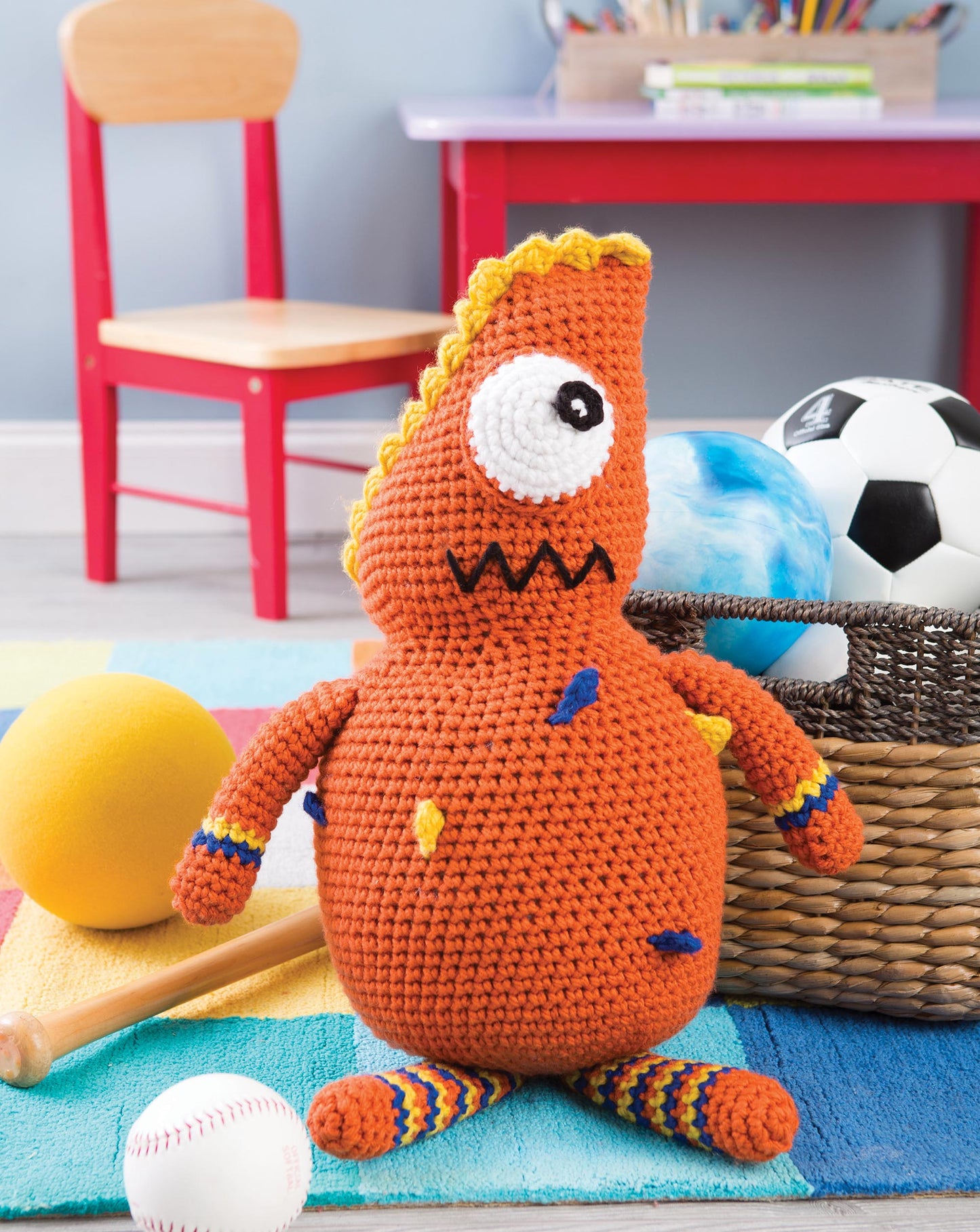 A Huggable Monsters plush from Leisure Arts, featuring one eye and a zigzag mouth, sits on a colorful rug near a basket of sports balls, a yellow ball, a bat, and with a red chair in the background.
