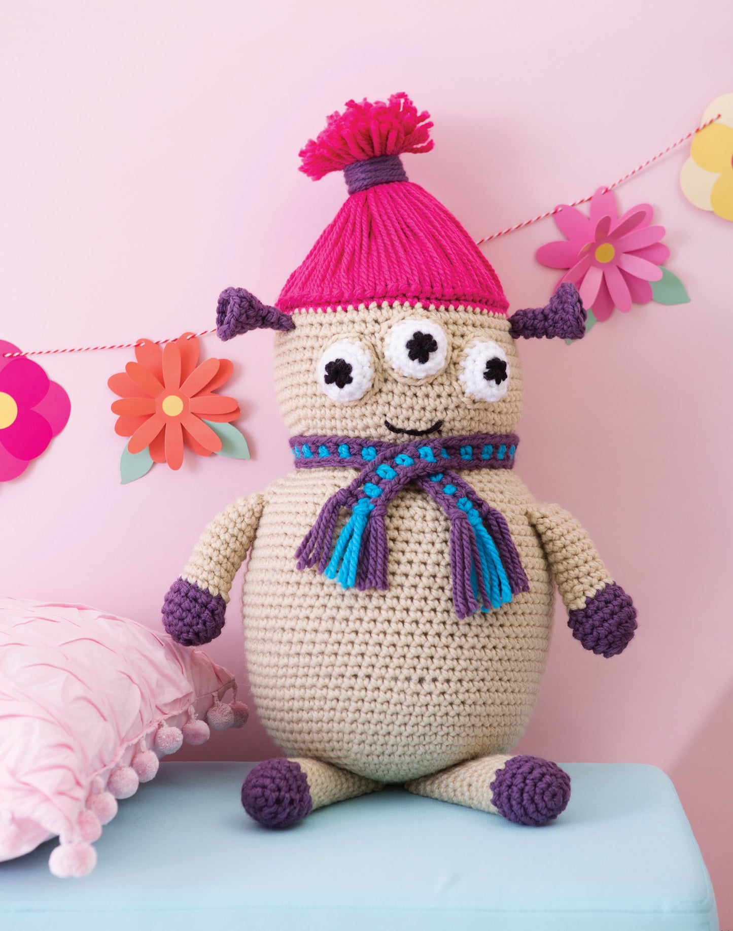 The Huggable Monsters doll by Leisure Arts features three eyes, purple hands, feet, and ears, a pink pompom hat, and a blue-purple scarf. Colorful paper flowers hang in the background. Ideal for fans or beginner crocheters seeking inspiration.