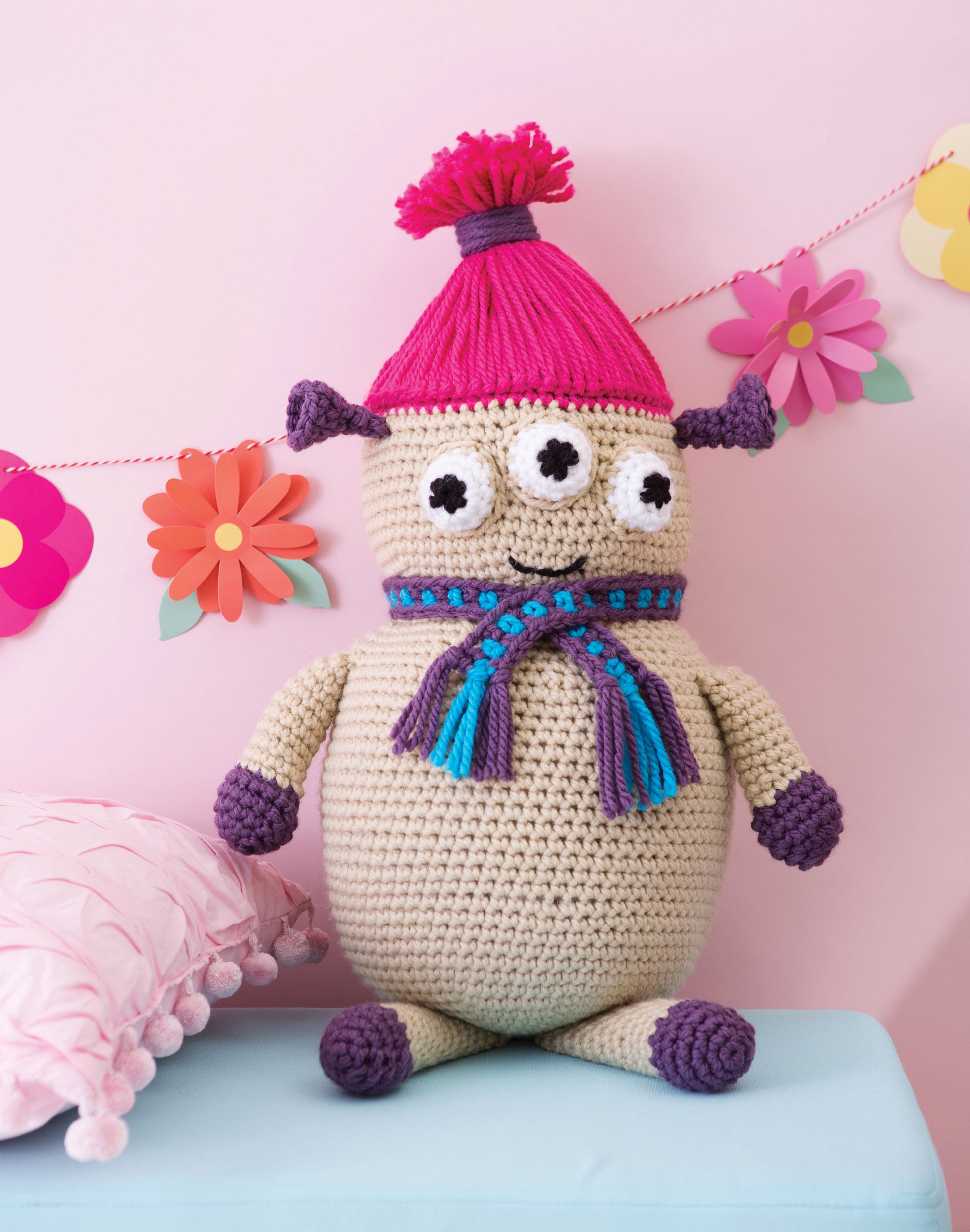 The Huggable Monsters doll by Leisure Arts features three eyes, purple hands, feet, and ears, a pink pompom hat, and a blue-purple scarf. Colorful paper flowers hang in the background. Ideal for fans or beginner crocheters seeking inspiration.