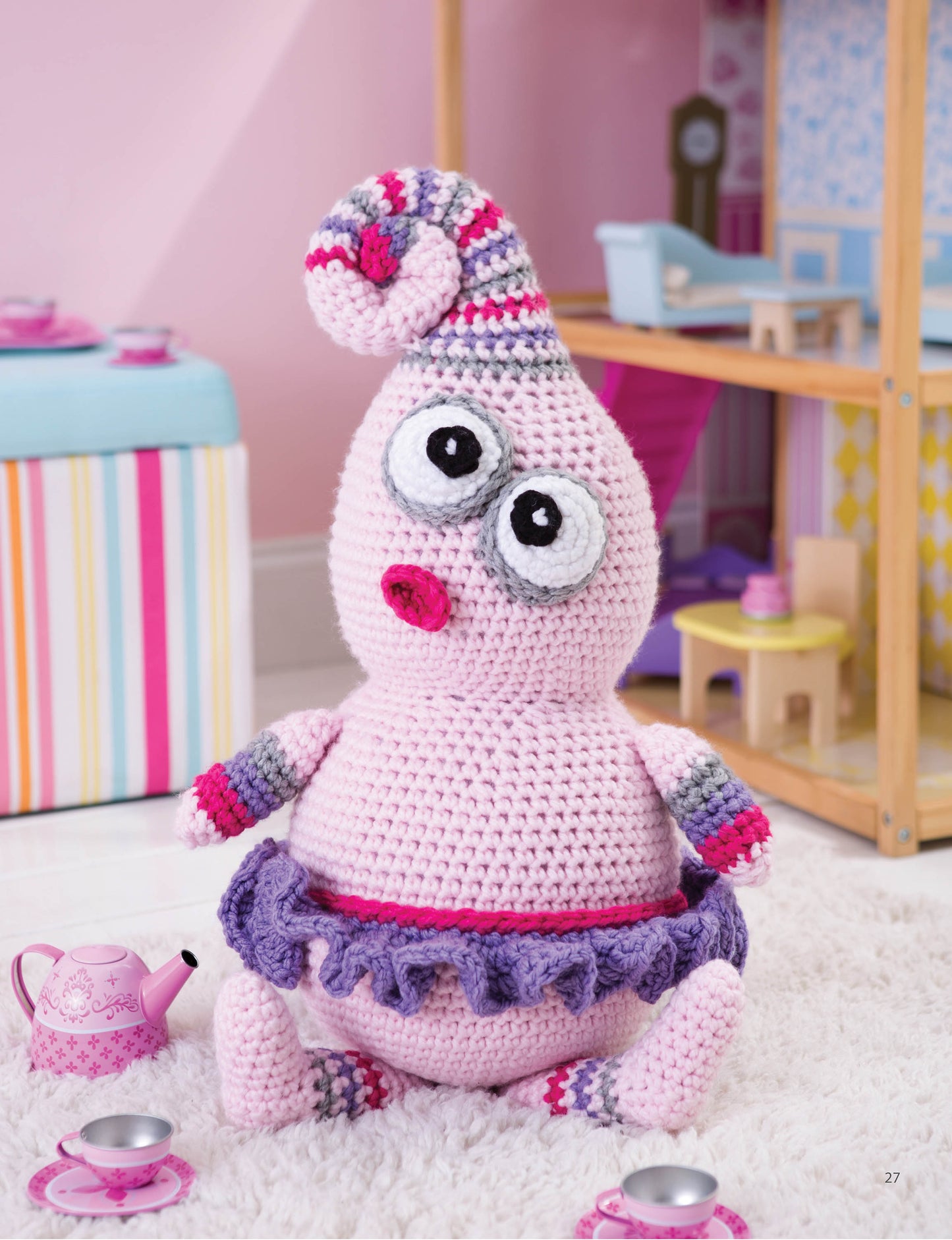 The Leisure Arts Huggable Monsters pink doll, with big eyes and beginner-friendly crochet design, sits near a dollhouse and toy tea set. Perfect for pastel playrooms, she inspires new monster patterns and adds charming decor.