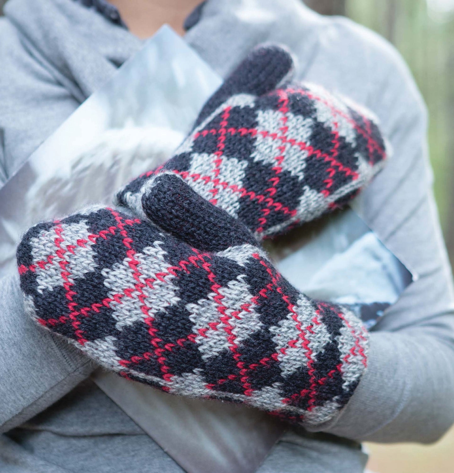A person in a light gray hoodie outdoors holds two photos while wearing Leisure Arts Fair Isle Mittens, featuring unique gray, black, and red checkered crochet patterns.