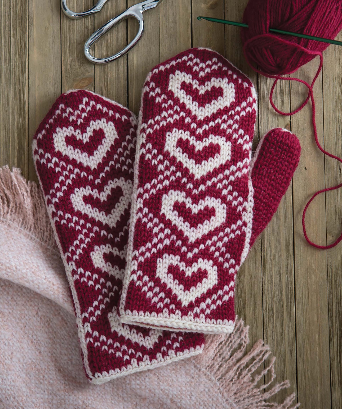 The Leisure Arts Fair Isle Mittens, featuring red and white heart patterns, rest on a light pink fringed blanket beside red yarn, knitting needles, and metal scissors on a wooden surface.