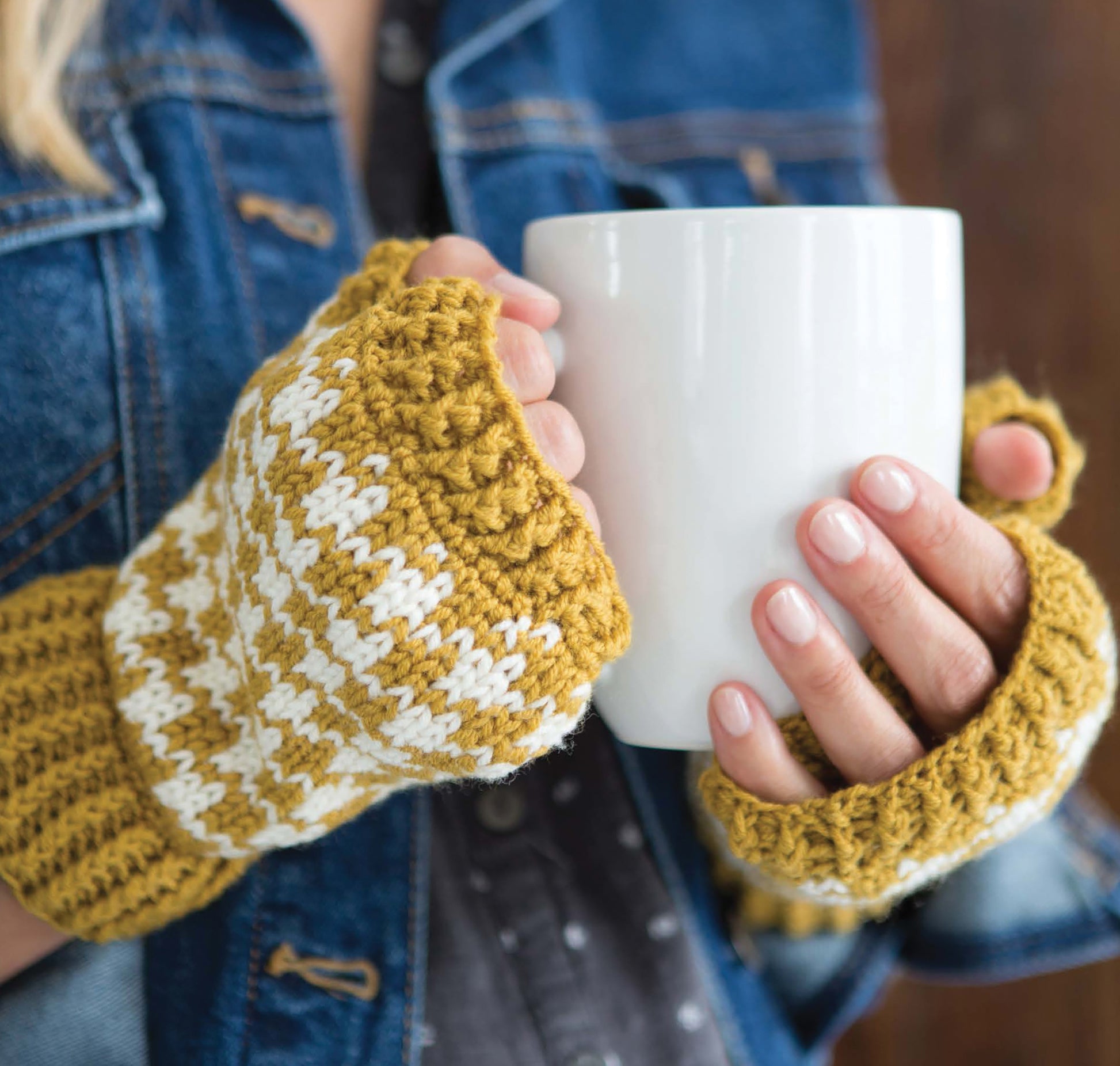 A person in a denim jacket wears Fair Isle Mittens by Leisure Arts and holds a large white mug with both hands.