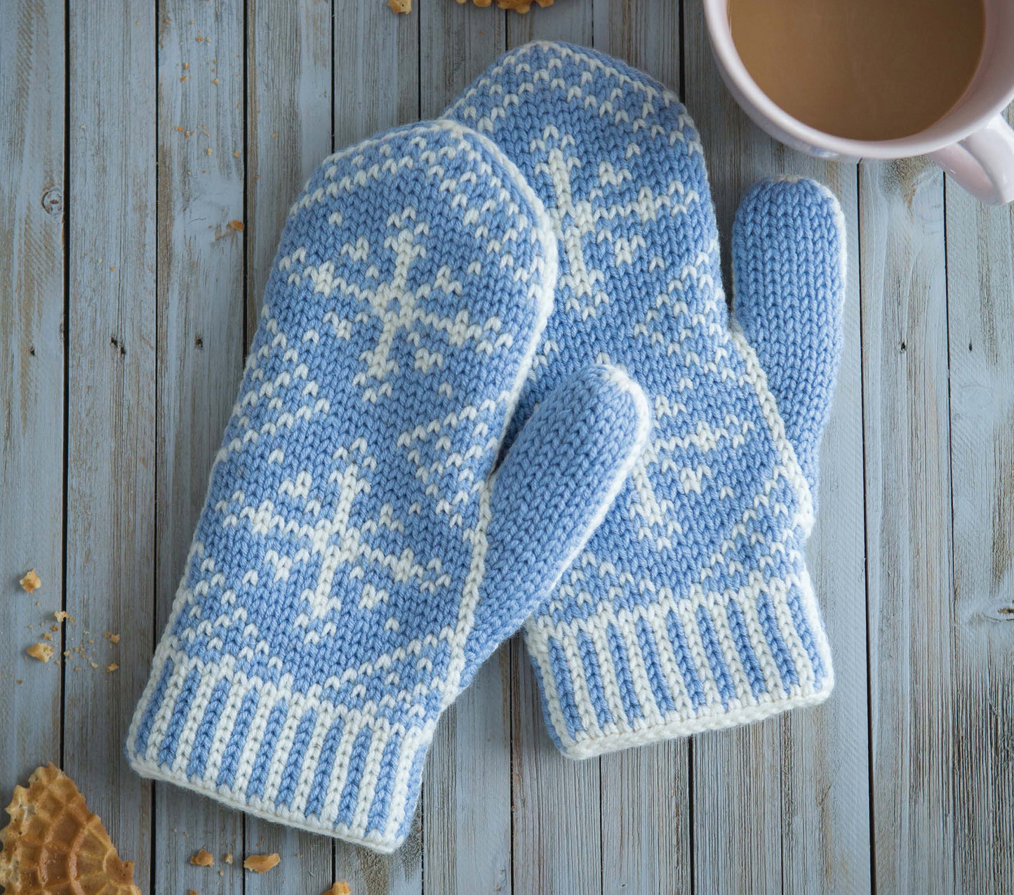 The Fair Isle Mittens by Leisure Arts, featuring blue and white crochet with snowflake patterns, rest on a light wooden surface beside a coffee cup and scattered waffle cookies.