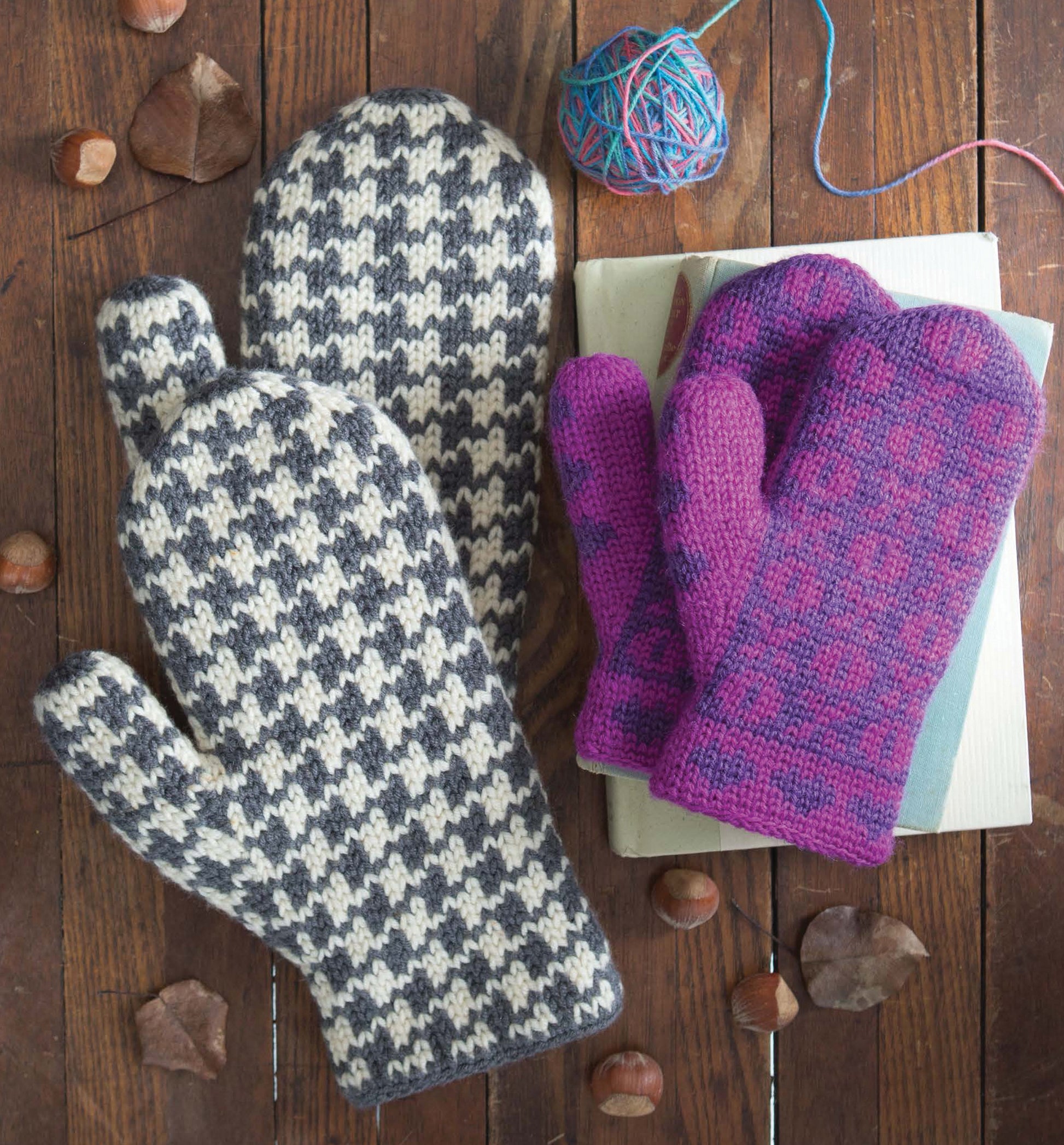 Two pairs of Leisure Arts Fair Isle Mittens—one black and white houndstooth, one purple and pink—are arranged on books and wood with chestnuts, a yarn ball, and charming mitten patterns on display.