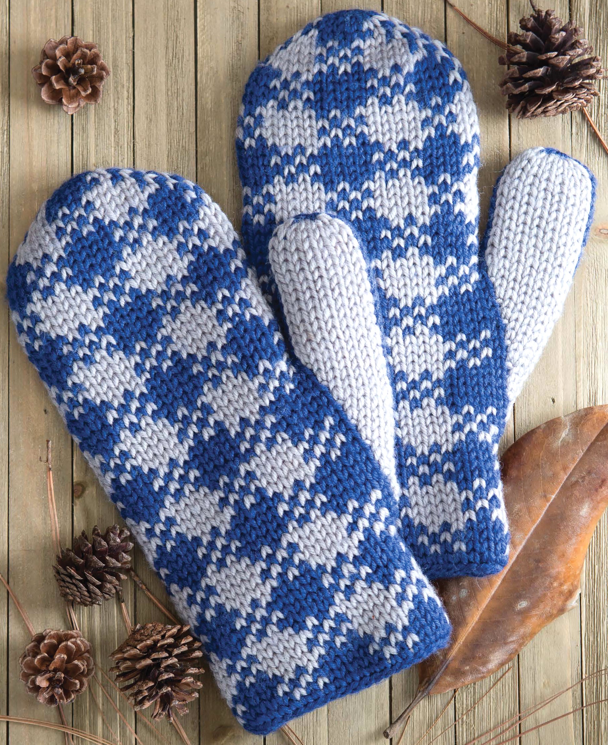 The Fair Isle Mittens by Leisure Arts feature a blue and white diamond crochet pattern, displayed on wood with pinecones and dried leaves for a cozy look.