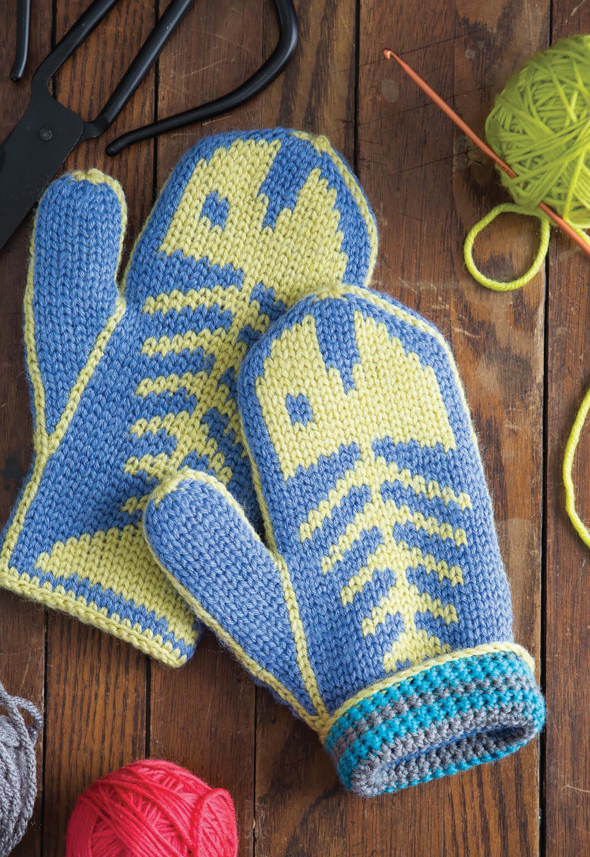 The Fair Isle Mittens by Leisure Arts feature blue and yellow fish skeleton designs and are displayed on wood with yarn, a crochet hook, and pliers—perfect inspiration for creating unique mitten patterns.