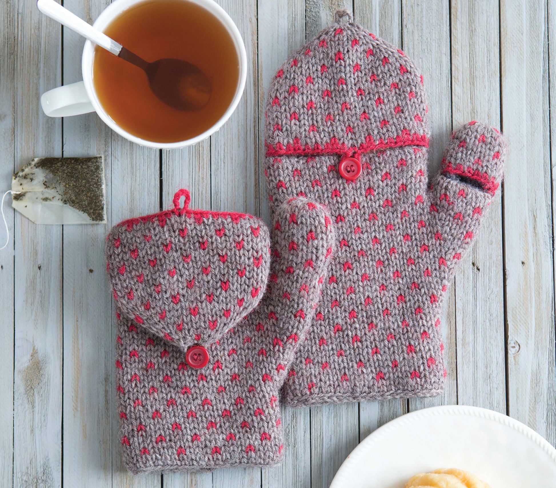 A cup of tea with a spoon and a tea bag sits beside two Leisure Arts Fair Isle Mittens with red dots and buttons on a light wooden surface; the edge of a white plate appears at the bottom right.