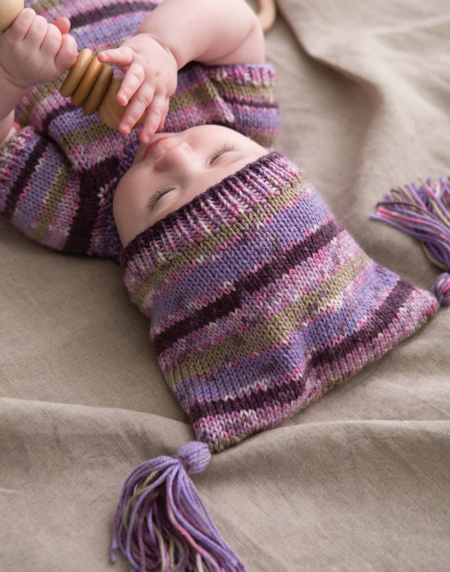 A baby in a striped purple, pink, and green knit outfit—made with medium weight yarn and seamless knits from Modern Baby Knitting by Leisure Arts—lies on a beige blanket holding a wooden rattle.