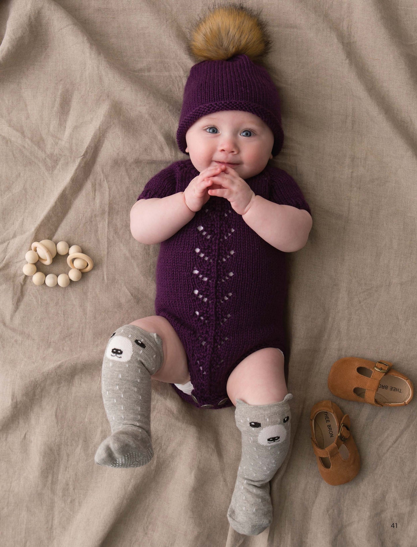 A baby wears a seamless purple knit outfit and pom-pom hat, made from medium weight yarn using Leisure Arts’ Modern Baby Knitting. The scene includes gray bear-face socks, brown shoes, and a wooden teether on a beige blanket.