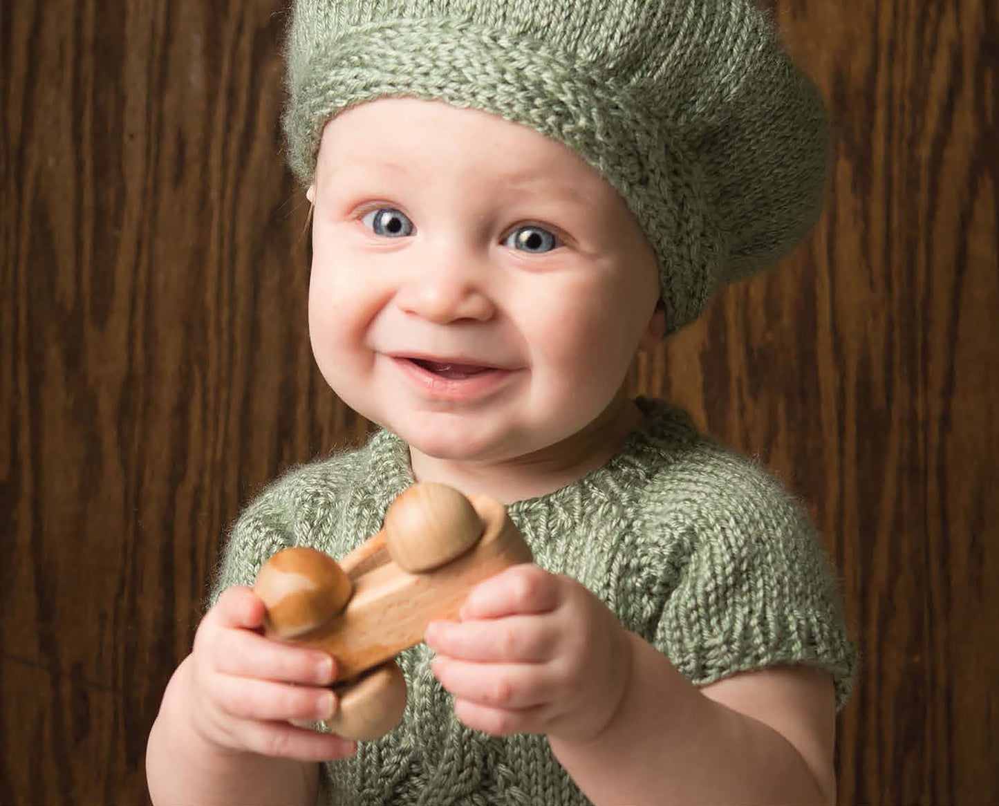 A smiling baby wears a green outfit and hat made from medium weight yarn, created with the Modern Baby Knitting pattern by Leisure Arts, holding a wooden toy car against a brown wooden background.