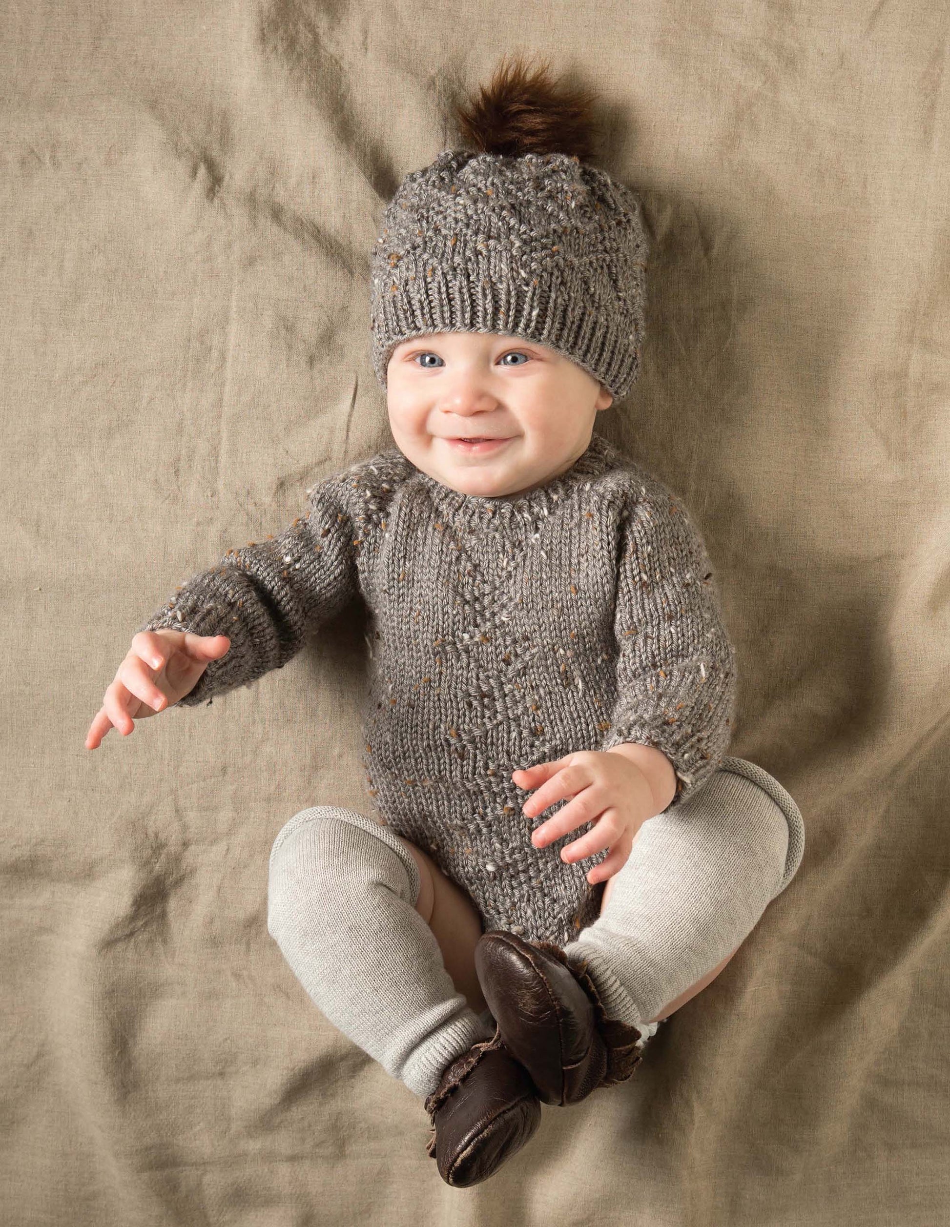 A baby in a gray knitted sweater and matching pom-pom hat from Modern Baby Knitting by Leisure Arts sits on a beige fabric background, paired with light gray tights and brown shoes.