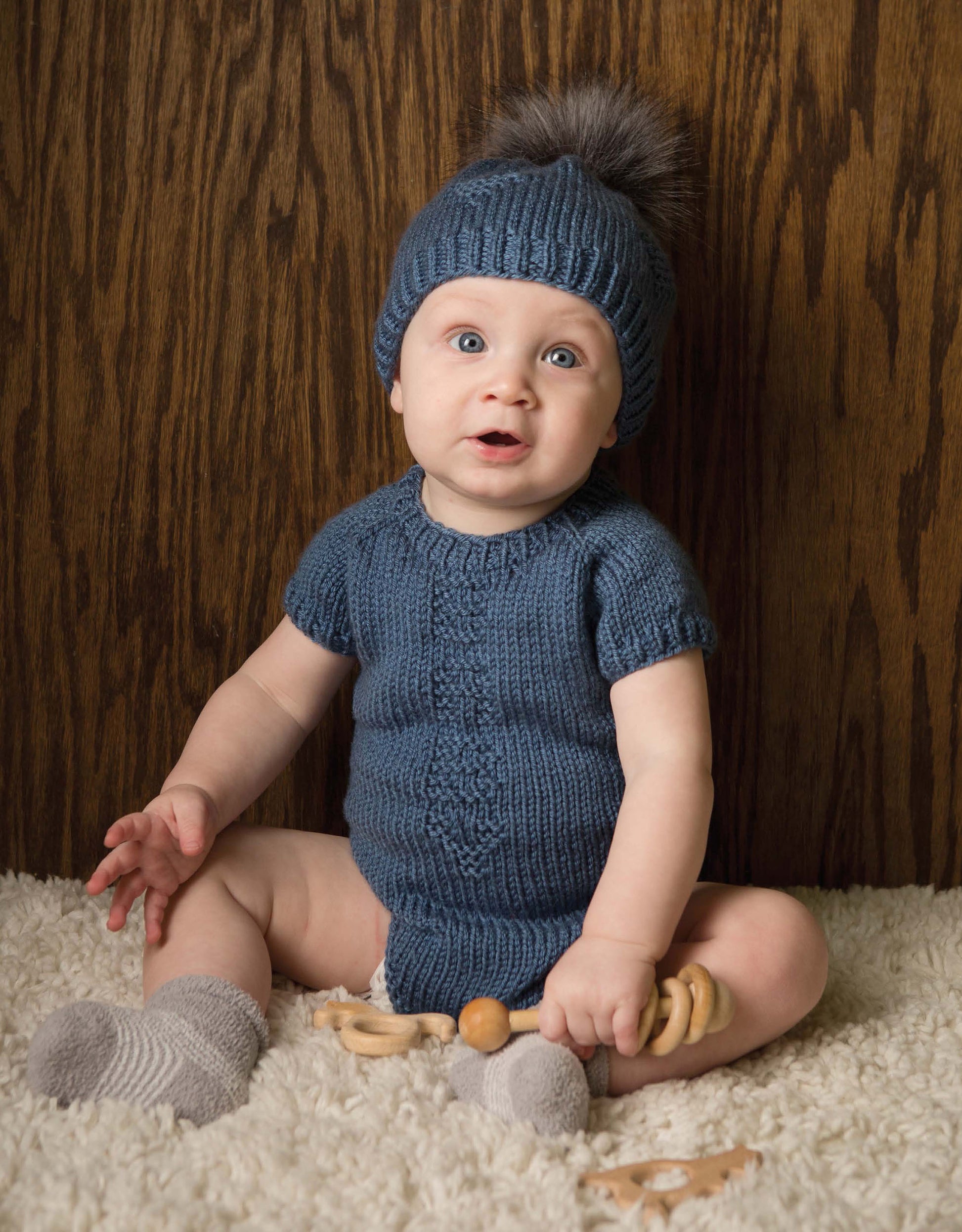 A baby in a blue knit outfit and pom-pom hat from Modern Baby Knitting by Leisure Arts sits on a fuzzy rug with wooden toys, against a wooden background.