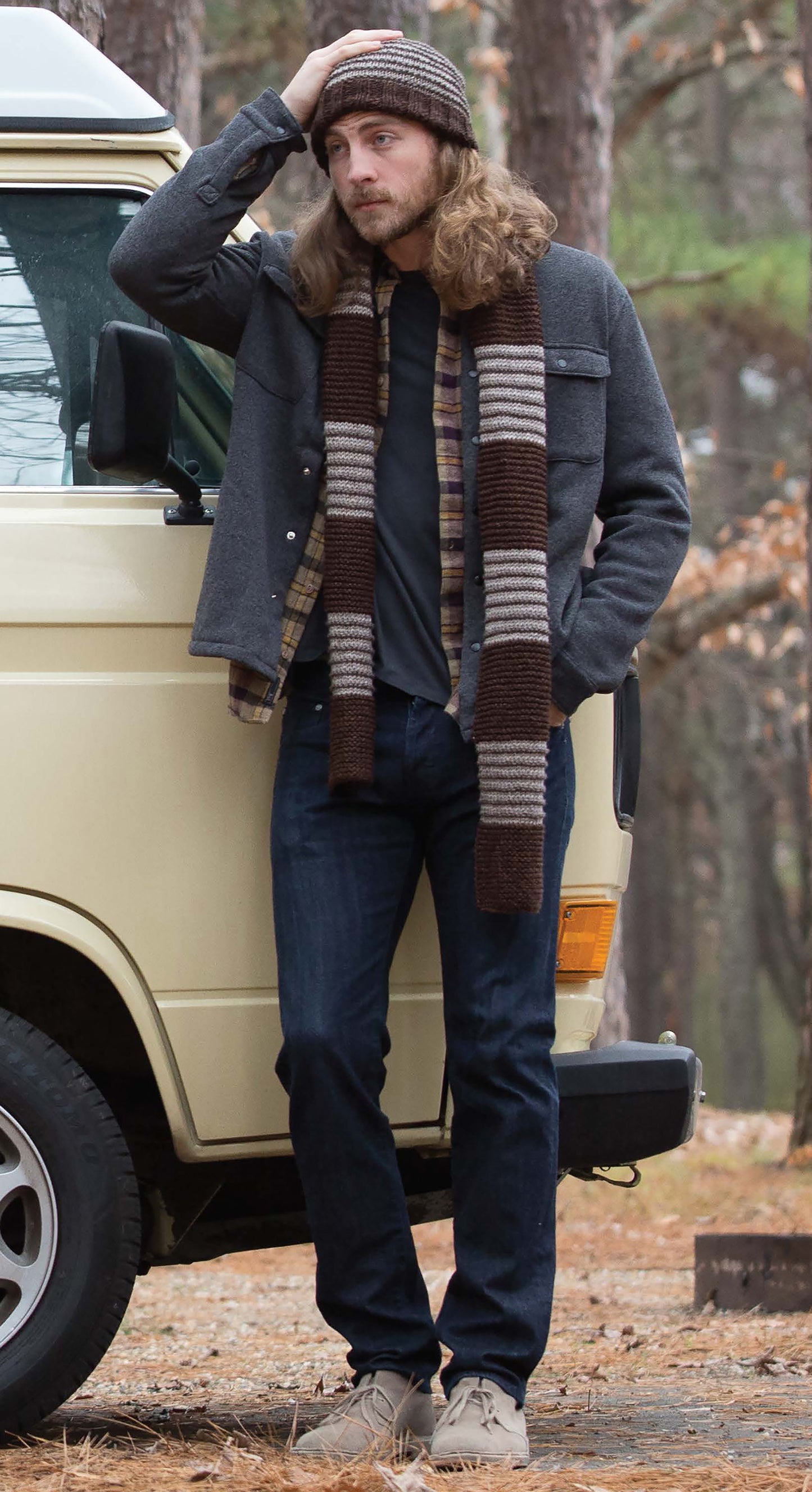 A man with long hair, wearing a knit hat from "Everyday Men's Knits" by Leisure Arts, plus a striped scarf and casual outfit, stands by a cream van in the woods, touching his hat and gazing into the distance.