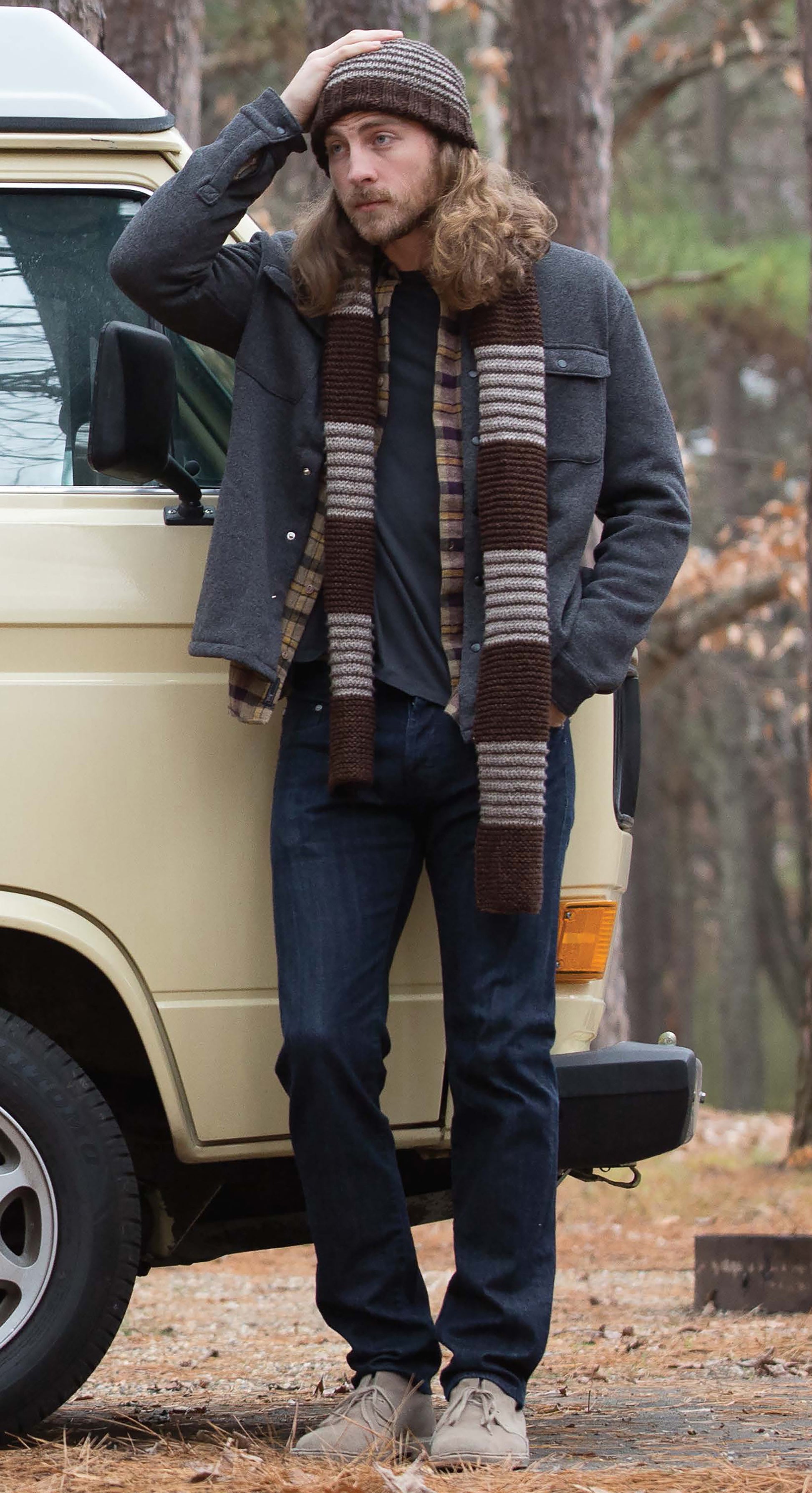 A man with long hair, wearing a knit hat from "Everyday Men's Knits" by Leisure Arts, plus a striped scarf and casual outfit, stands by a cream van in the woods, touching his hat and gazing into the distance.