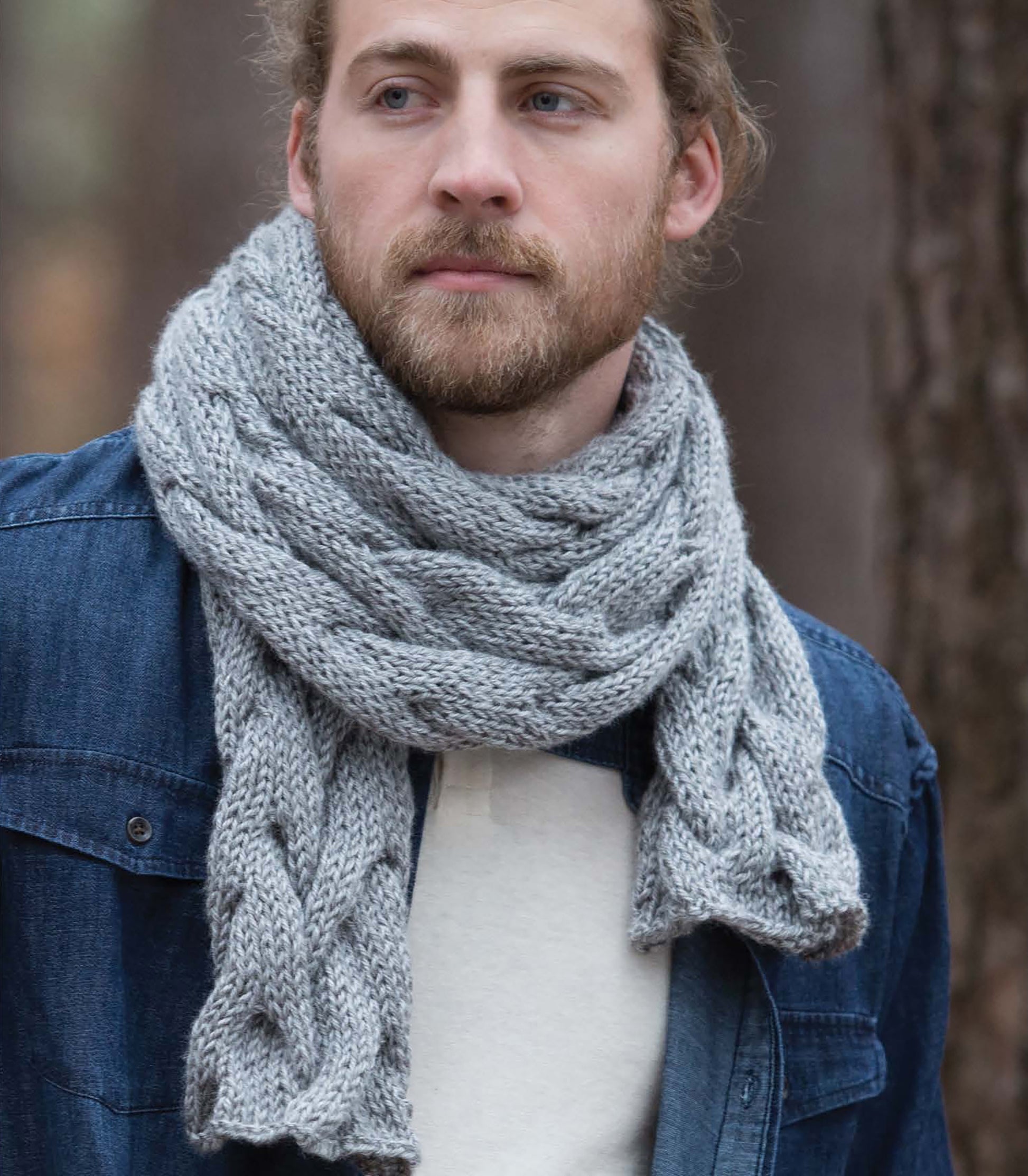 A man with a beard and long hair models the thick, braided gray scarf from Leisure Arts' "Everyday Men's Knits," worn over a blue denim jacket and white shirt while standing among trees outdoors.