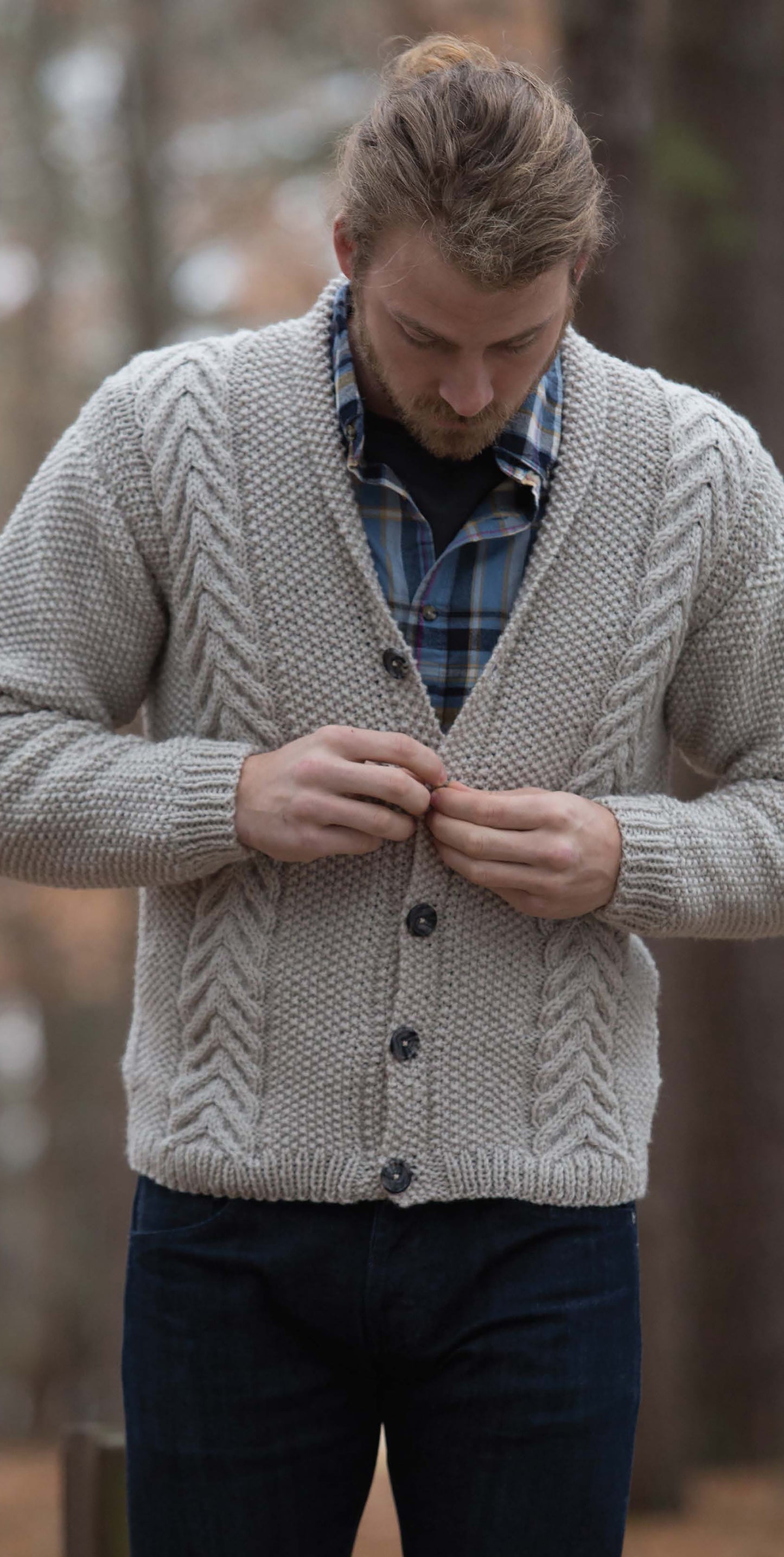 A bearded man buttons a chunky light gray cable-knit cardigan over a blue plaid shirt—an inviting look from Leisure Arts' Everyday Men's Knits. A blurred, earthy background hints at an outdoor, wooded setting.
