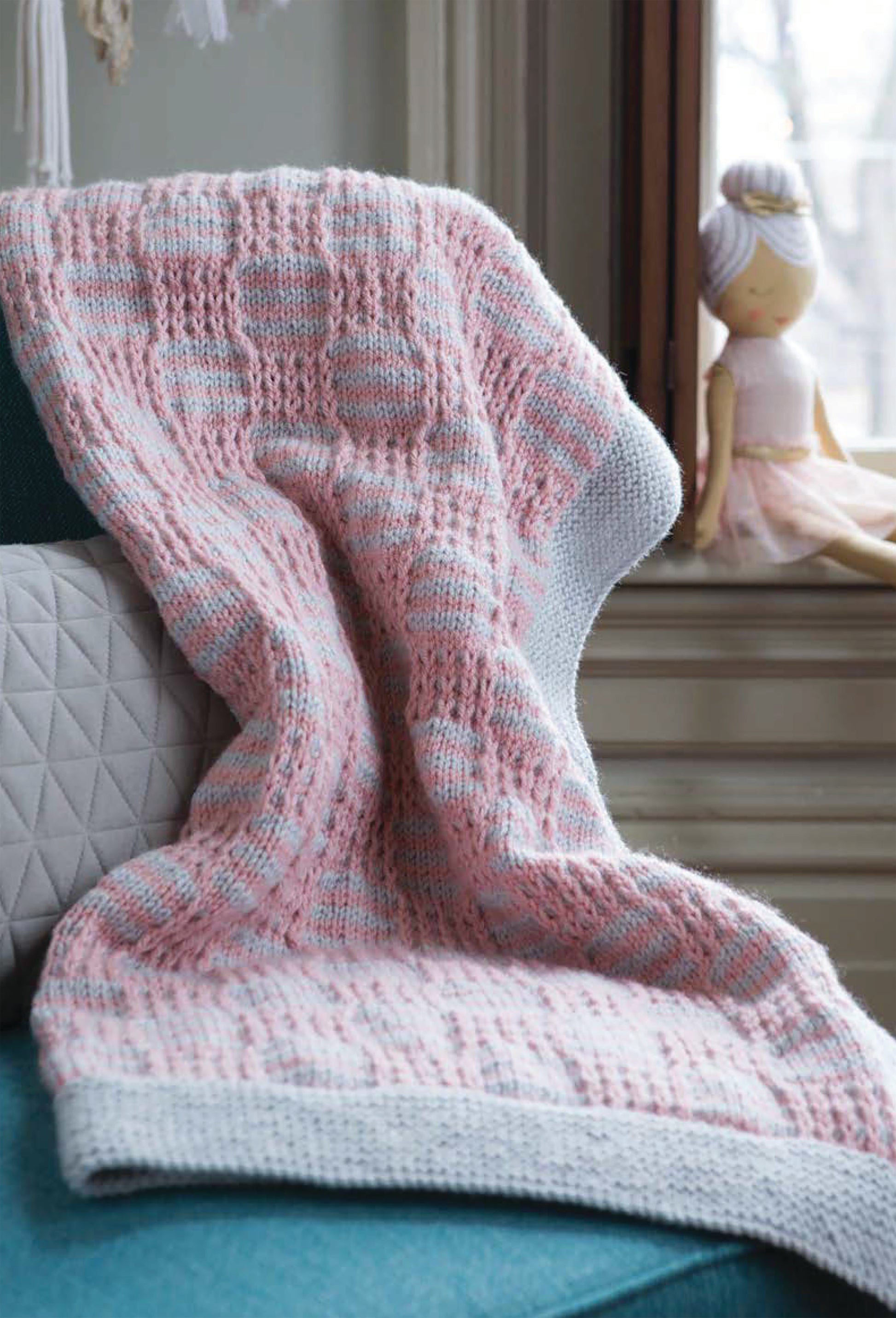 A cozy knit blanket from "The Beginner's Guide to Mosaic Knitting" by Leisure Arts is spread over a blue couch with a gray cushion, while a cloth doll in pink sits on a windowsill in the background.