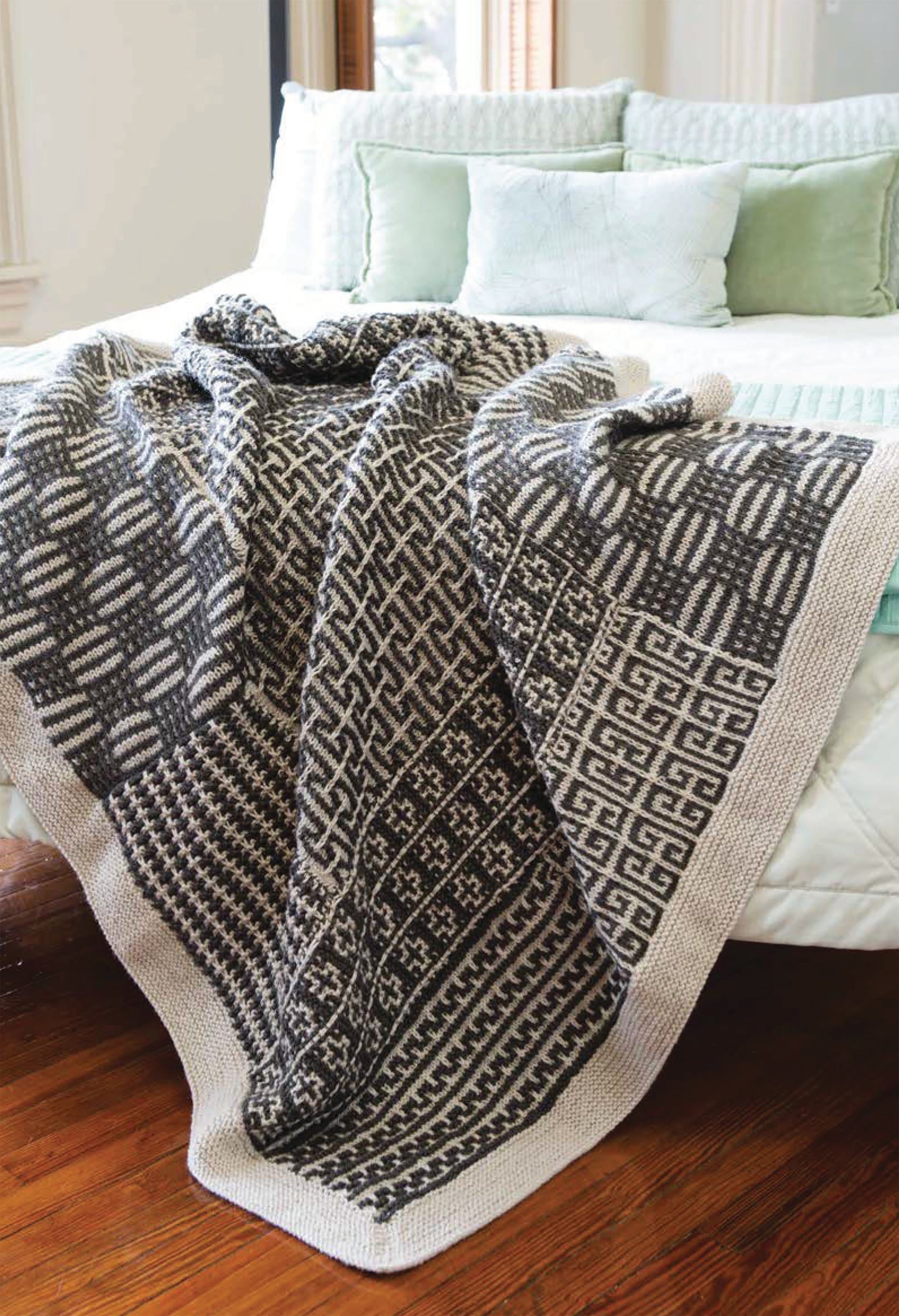 The Beginner's Guide to Mosaic Knitting by Leisure Arts features gray and white mosaic knit patterns, perfect for beginners. Displayed draped over a bed with light green and white pillows for a stylish touch.