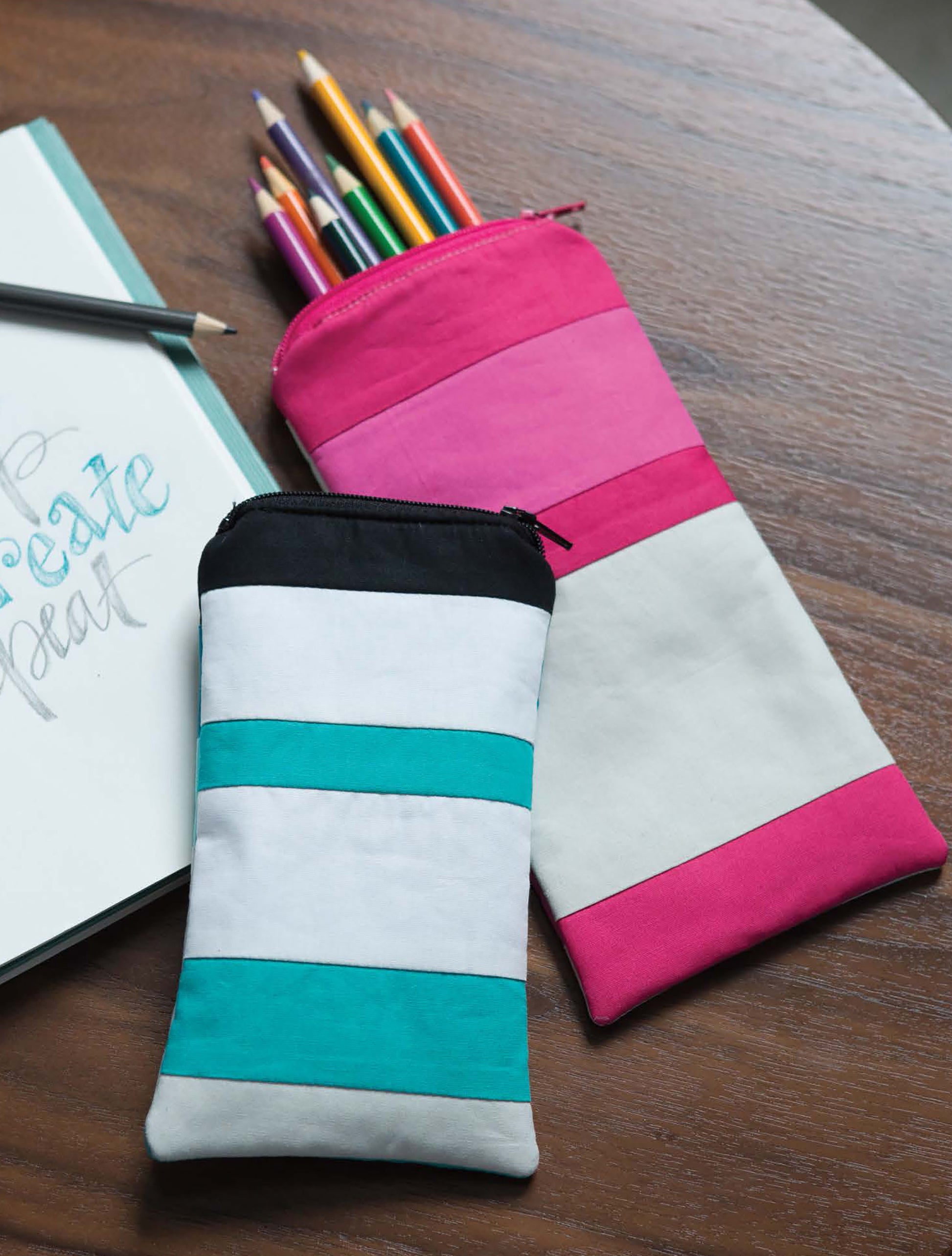 Two striped patchwork pouches, featured in the "Modern Patchwork Digital Download" by Leisure Arts, rest on a wooden table—one pink and white with colored pencils inside, the other teal, white, and black. A sketchbook and pencil accompany them.