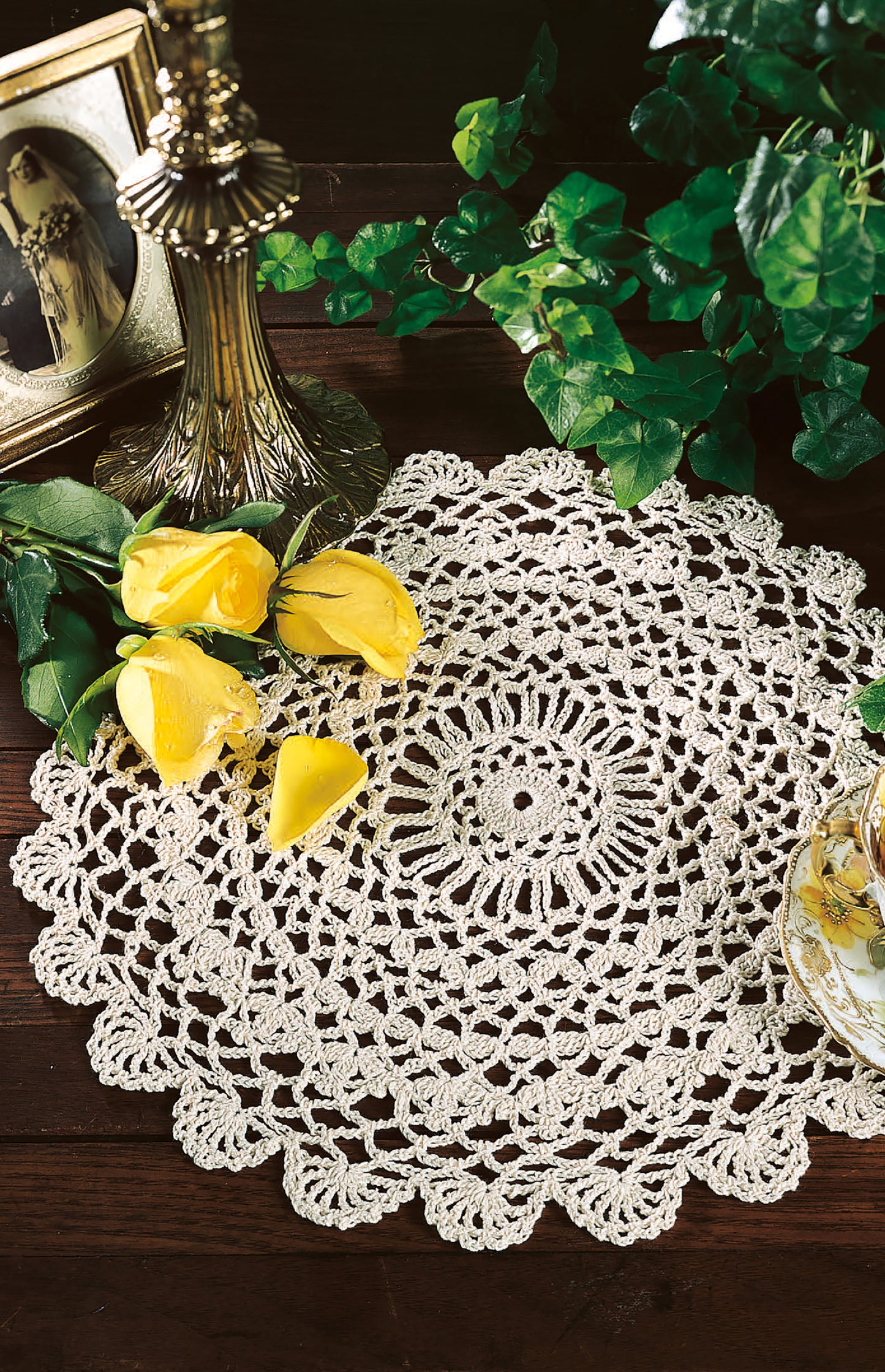 A copy of "Thread Crochet—A Beginner's Guide to 9 Projects" by Leisure Arts is displayed on a wooden table with yellow roses, a teacup, a vintage photo in a frame, a brass lamp, and green ivy leaves.