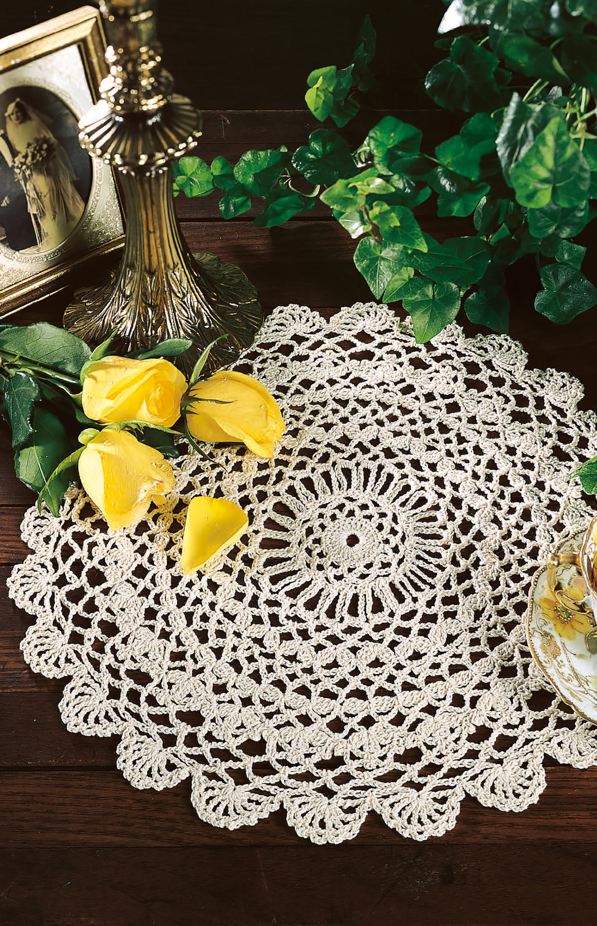 A copy of "Thread Crochet—A Beginner's Guide to 9 Projects" by Leisure Arts is displayed on a wooden table with yellow roses, a teacup, a vintage photo in a frame, a brass lamp, and green ivy leaves.