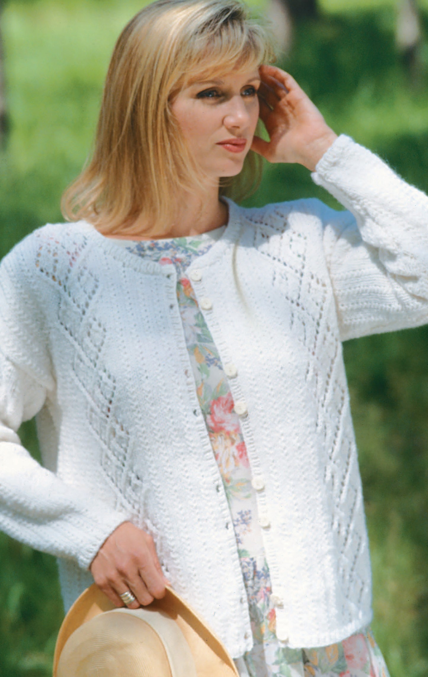 A woman in a white knit cardigan, ideal for beginner knitters using Leisure Arts' "Beginner's Guide Knit Stitches & Easy Projects," stands outside over a floral dress, holding a straw hat with greenery blurred in the background.
