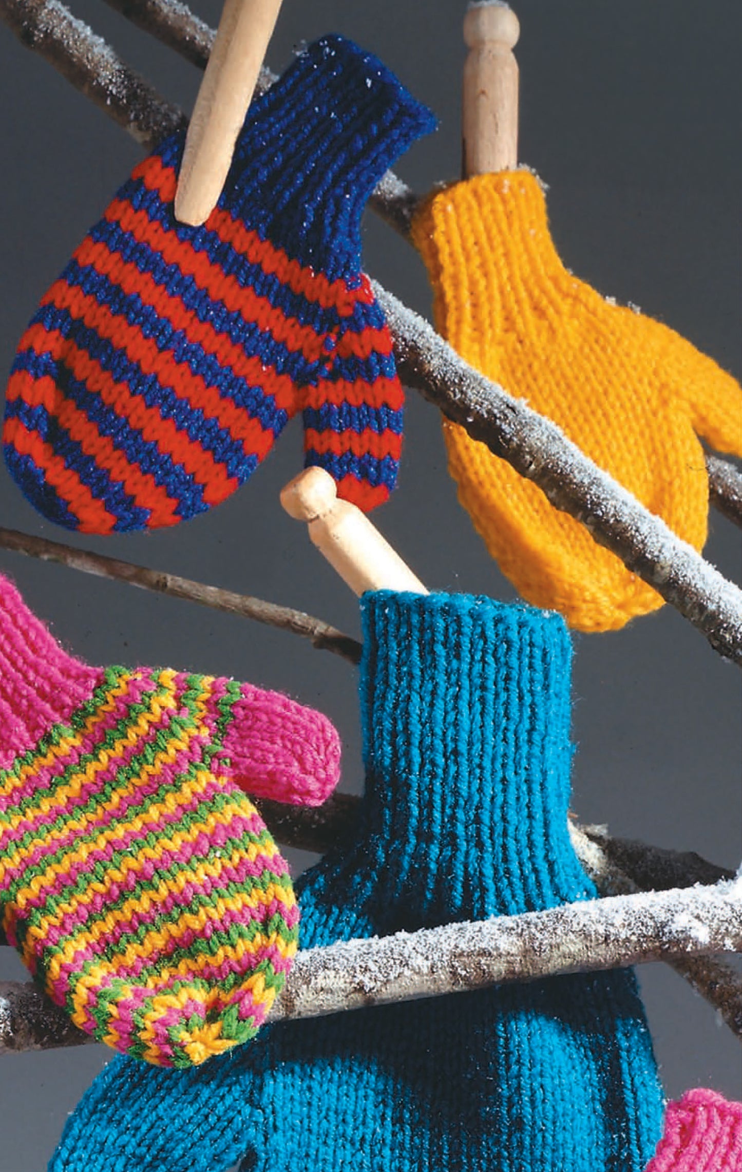 Four vibrant knit mittens—great for practicing stitches from Leisure Arts’ Beginner's Guide Knit Stitches & Easy Projects—are clipped to frosty branches: red and blue stripes, yellow, pink and green stripes, and solid blue.