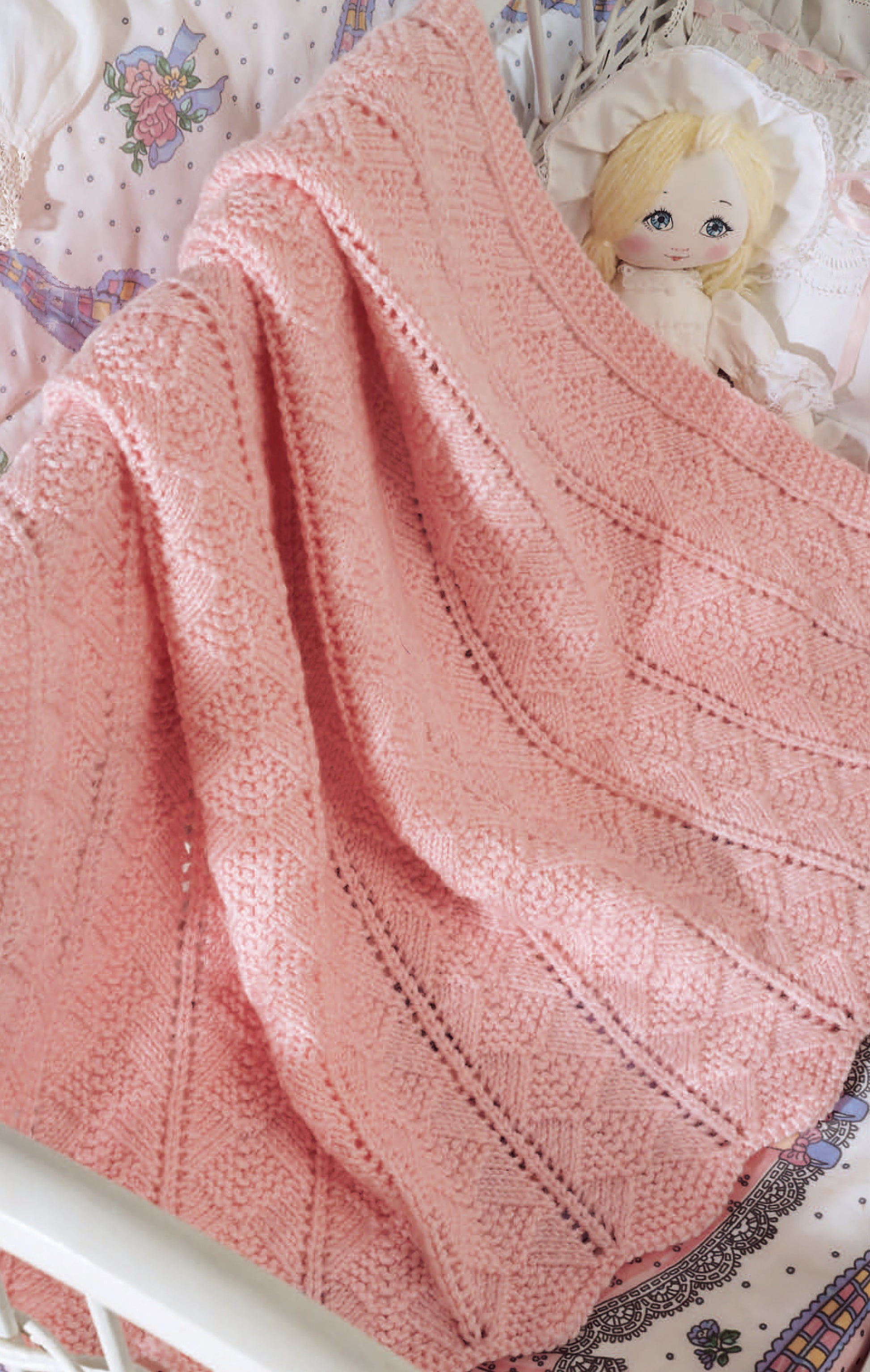 A blonde-haired doll rests in a crib, covered by a soft pink blanket crafted with knit stitches inspired by Leisure Arts' "Beginner's Guide Knit Stitches & Easy Projects," atop whimsical unicorn and star bedding with lace accents.
