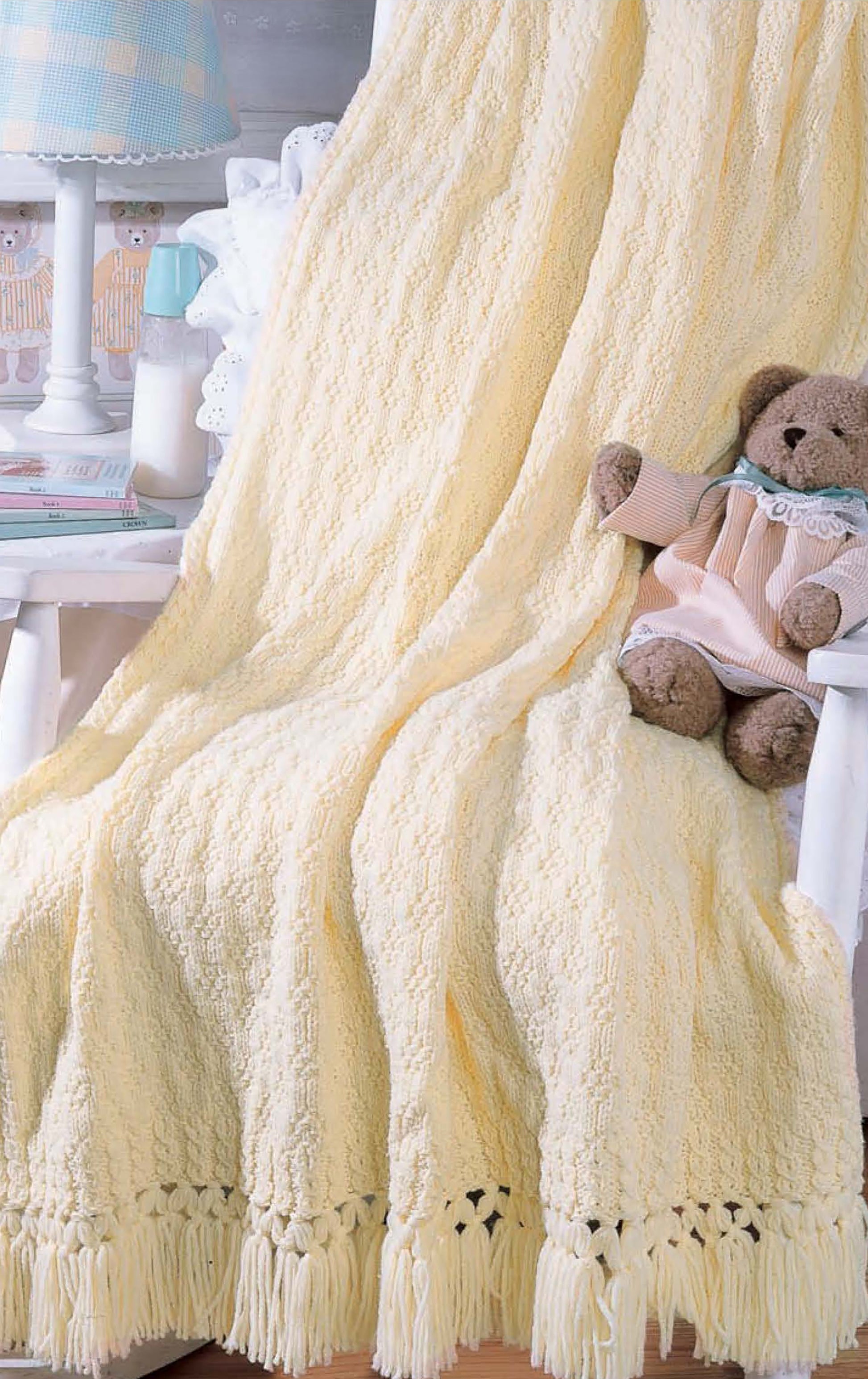 The cozy Snuggletime Baby Afghans by Leisure Arts, shown in soft yellow with fringe, is draped over a white chair beside a teddy bear and table in a warmly lit room—perfect for knit and crochet lovers.