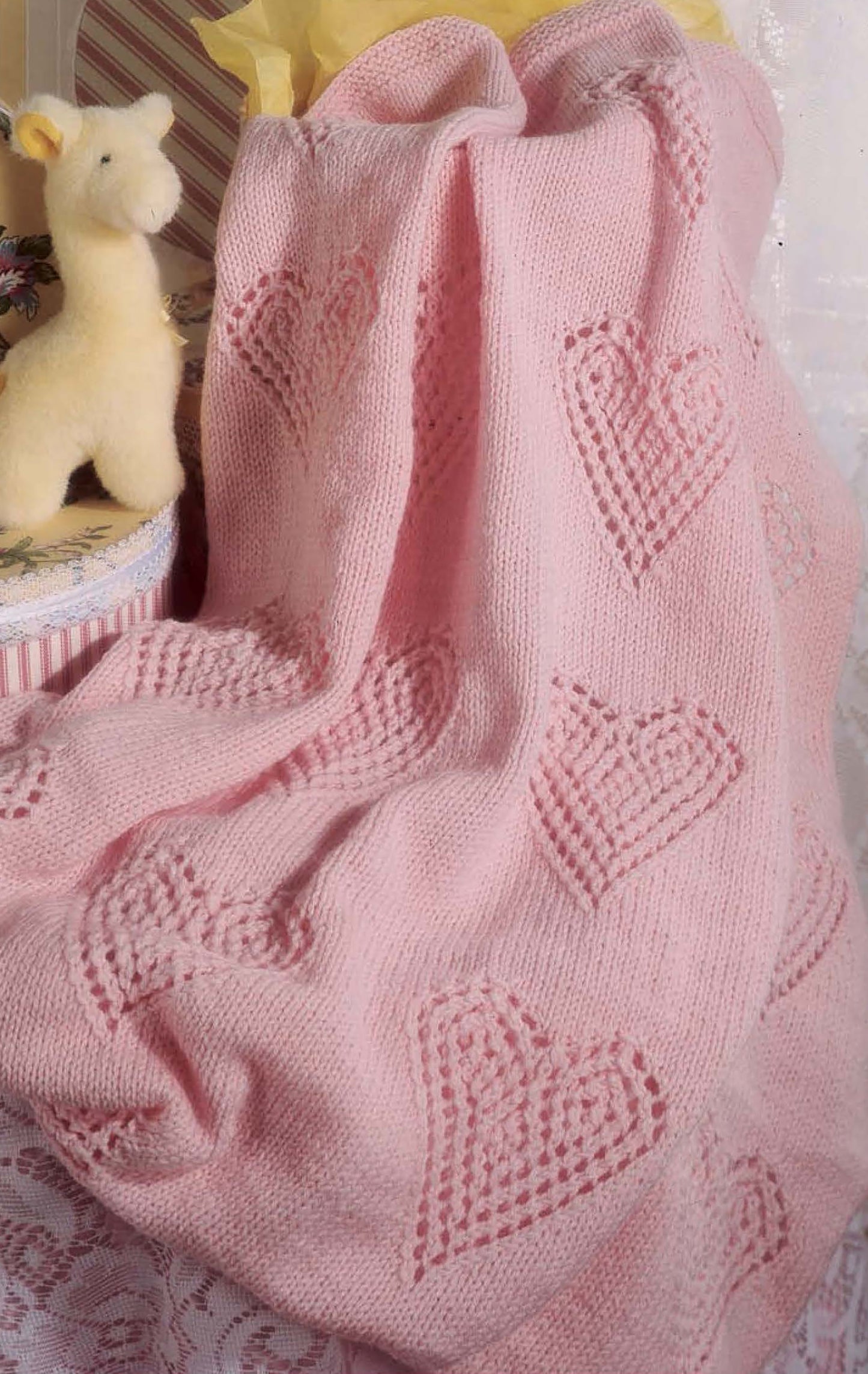 The "Snuggletime Baby Afghans" by Leisure Arts is a soft pink knitted blanket with a heart pattern, shown draped beside a plush giraffe—ideal for knit and crochet baby throws.