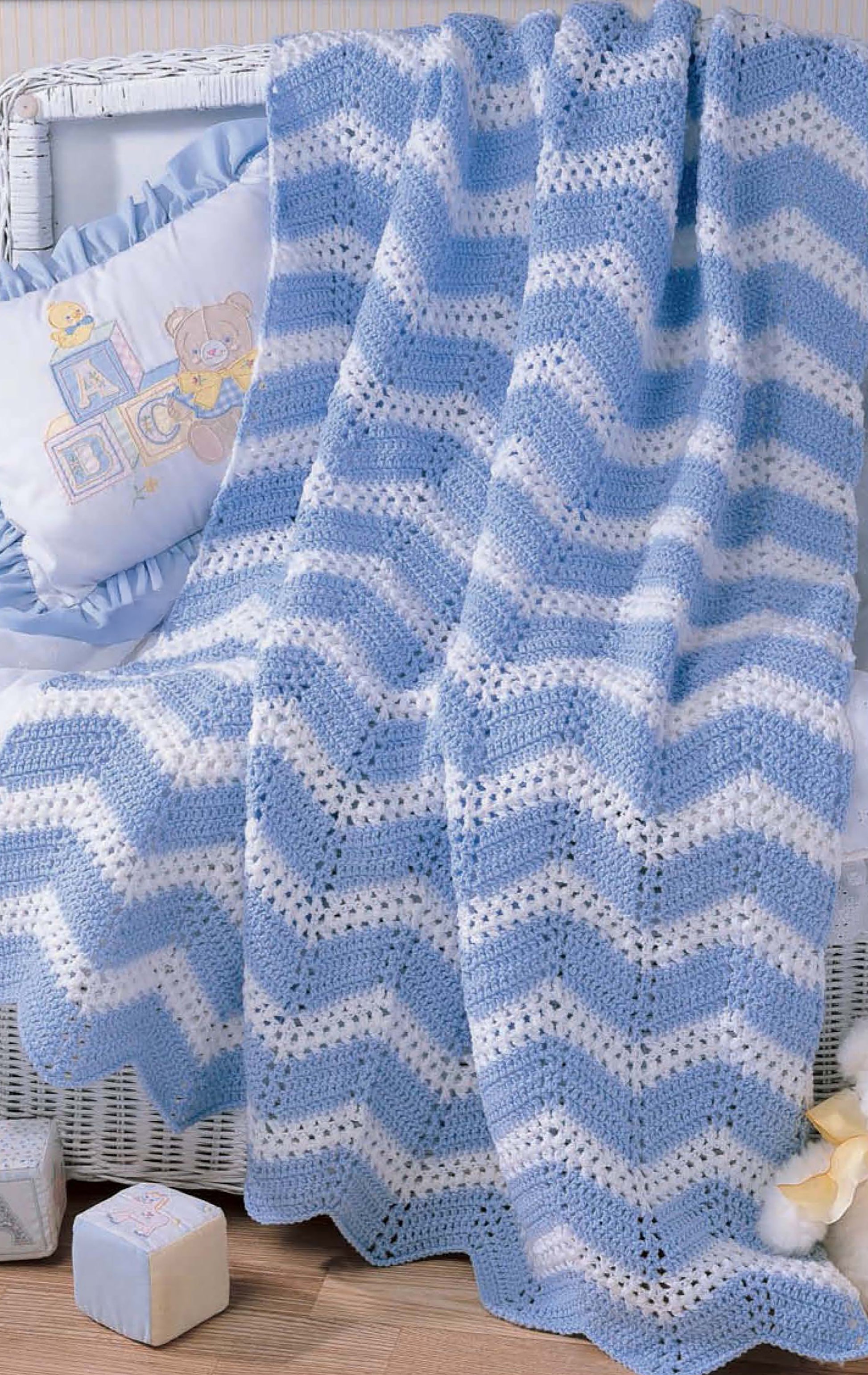 The Snuggletime Baby Afghans by Leisure Arts, featuring a blue and white chevron knit and crochet design, is draped over a white wicker crib with baby pillows and blocks arranged nearby on a wooden floor.