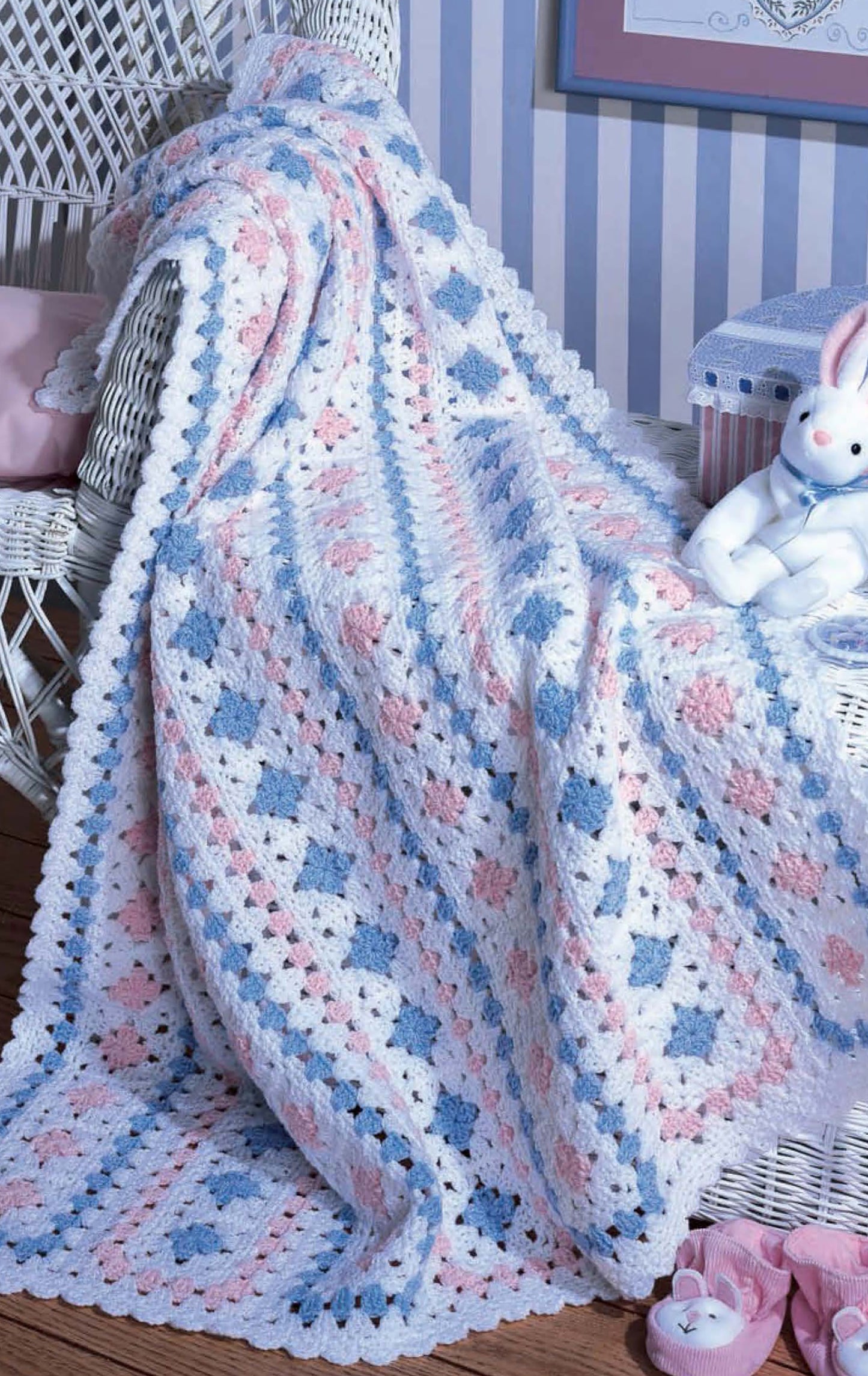 A Leisure Arts Snuggletime Baby Afghans throw with a white, pink, and blue granny square pattern drapes over a white wicker chair in the nursery, accompanied by a stuffed rabbit and striped storage box for added charm.