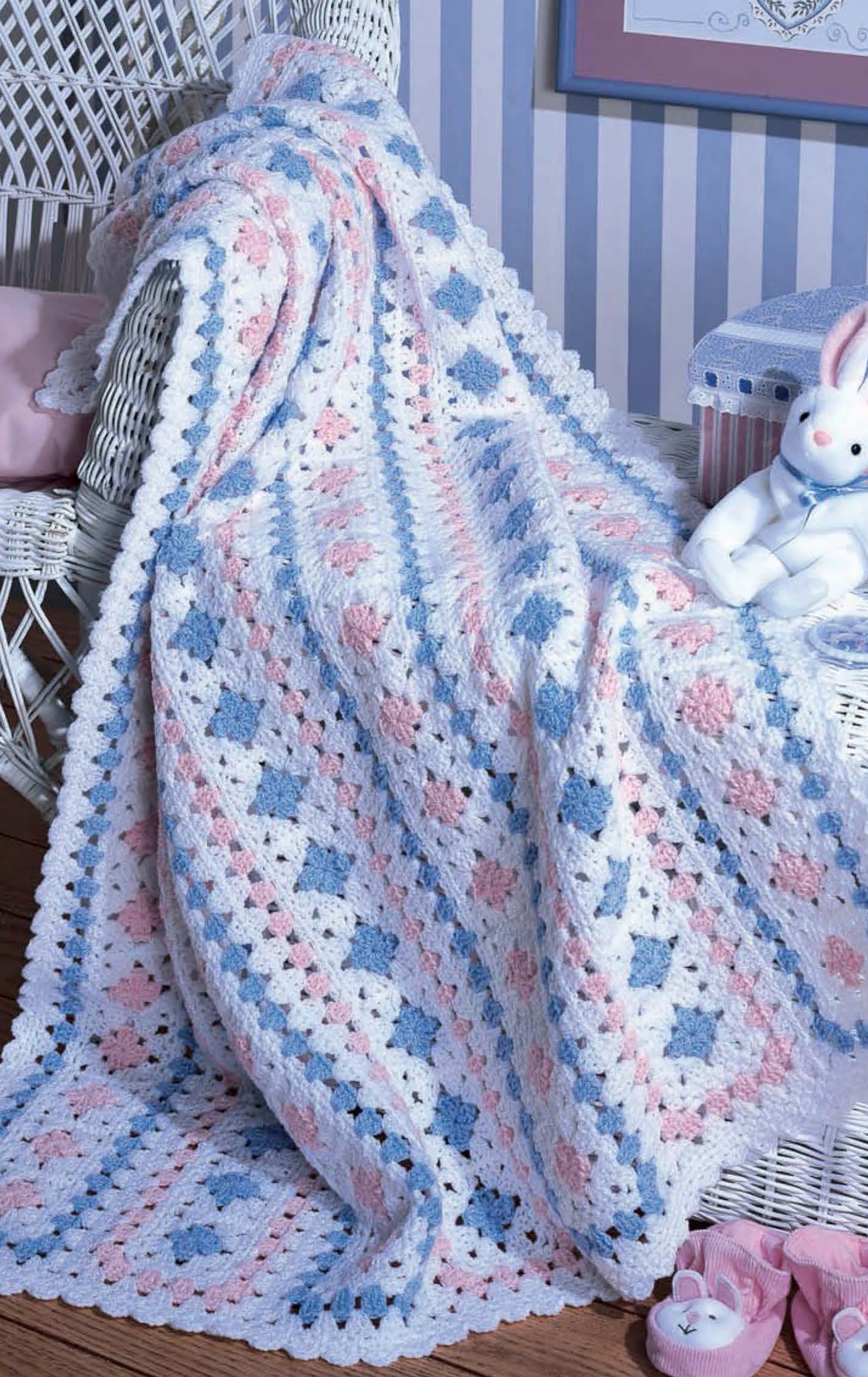 A Leisure Arts Snuggletime Baby Afghans throw with a white, pink, and blue granny square pattern drapes over a white wicker chair in the nursery, accompanied by a stuffed rabbit and striped storage box for added charm.