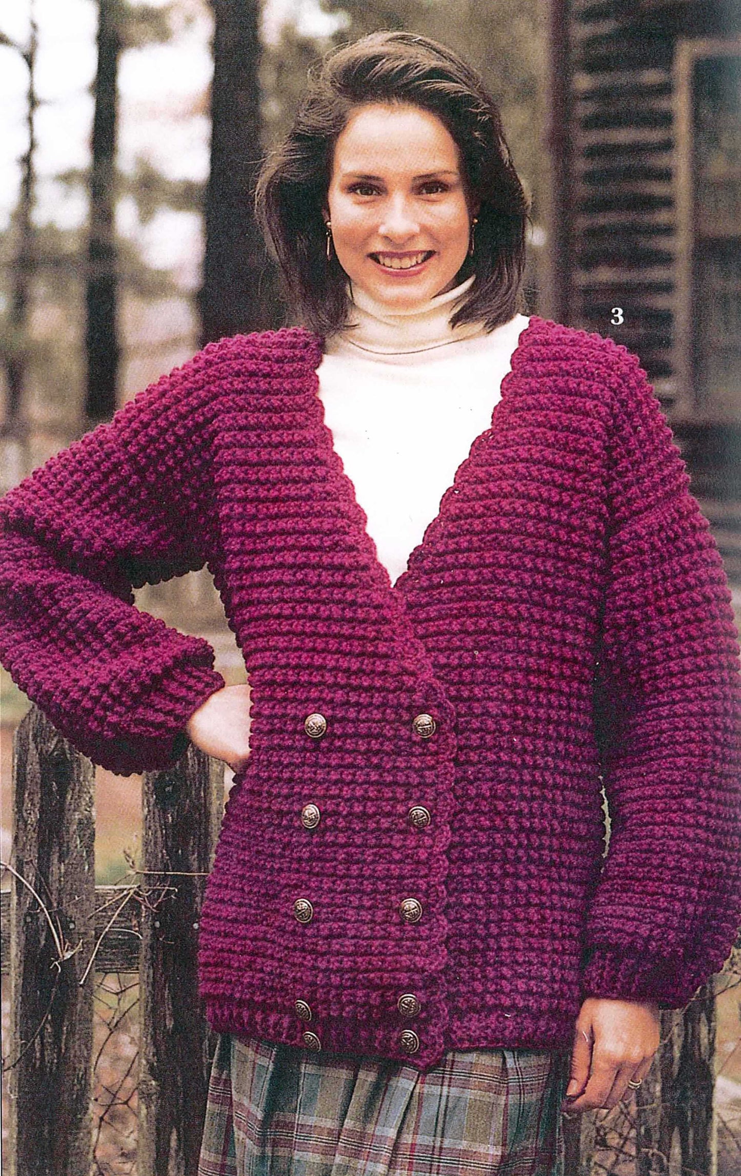 A woman stands outdoors by a wooden fence, smiling and wearing a deep purple double-breasted cardigan made using patterns from Leisure Arts’ “19 Crochet Stitches & Easy Projects” digital download, over a white turtleneck and plaid skirt.