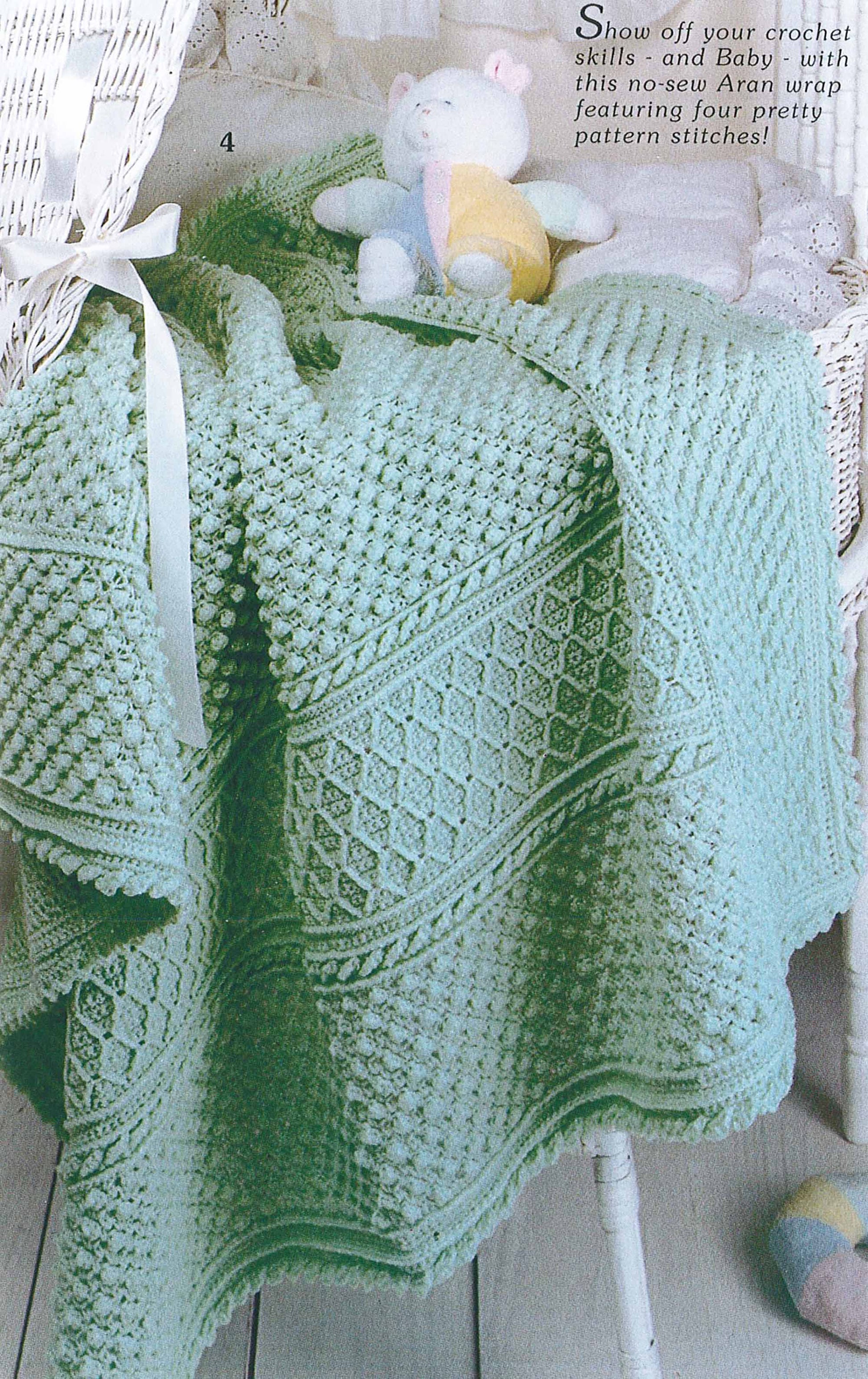 A soft, light green baby blanket made with textured stitches from Leisure Arts' "19 Crochet Stitches & Easy Projects" digital download is draped over a white wicker chair, paired with a pastel plush toy bear—perfect for beginner crocheters.