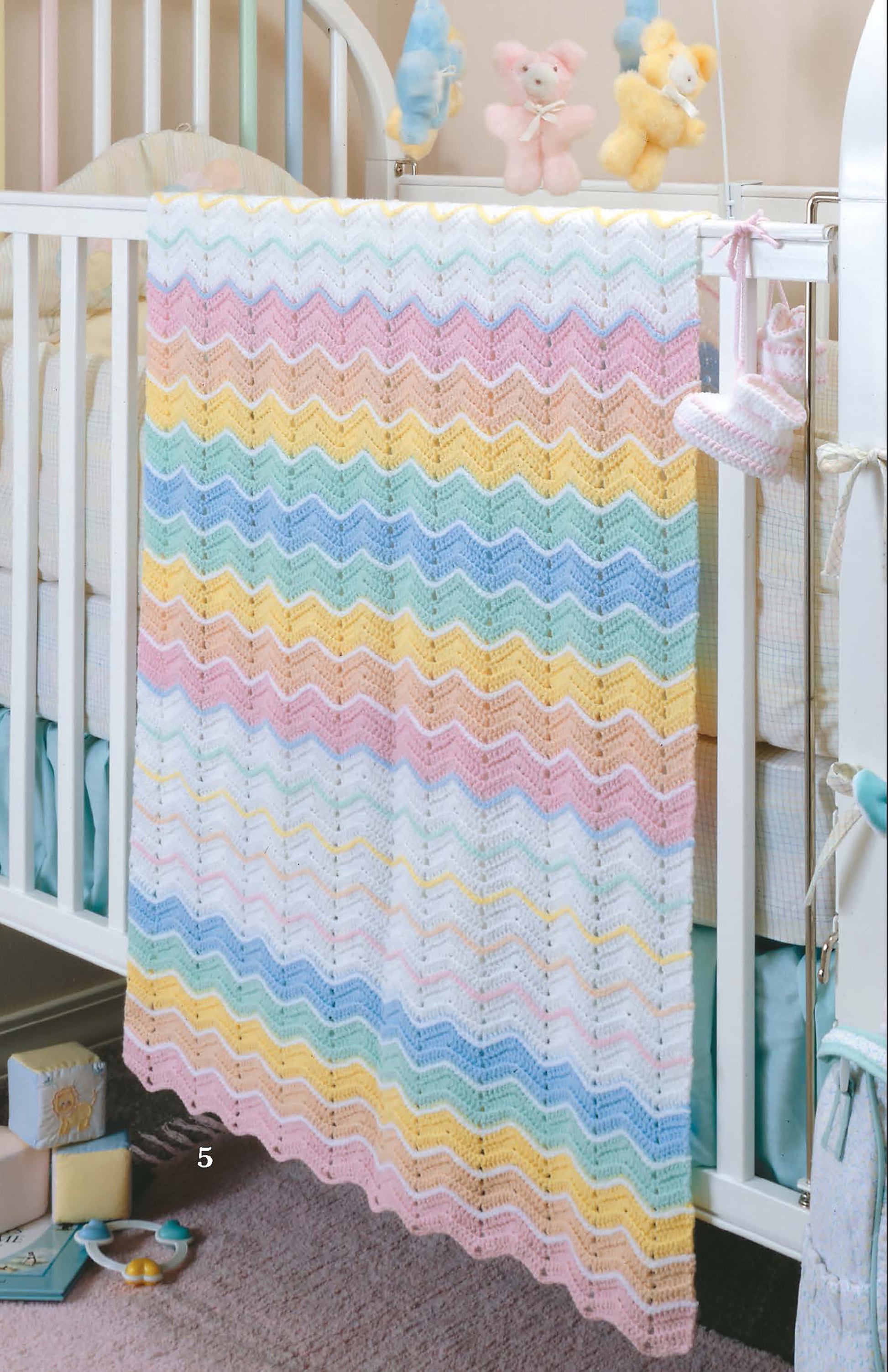 The Leisure Arts "Ripples for Baby to Crochet" pastel chevron-patterned blanket hangs over a white crib with plush bears, a pacifier, and pink booties. Soft blue and yellow accents brighten the nursery.